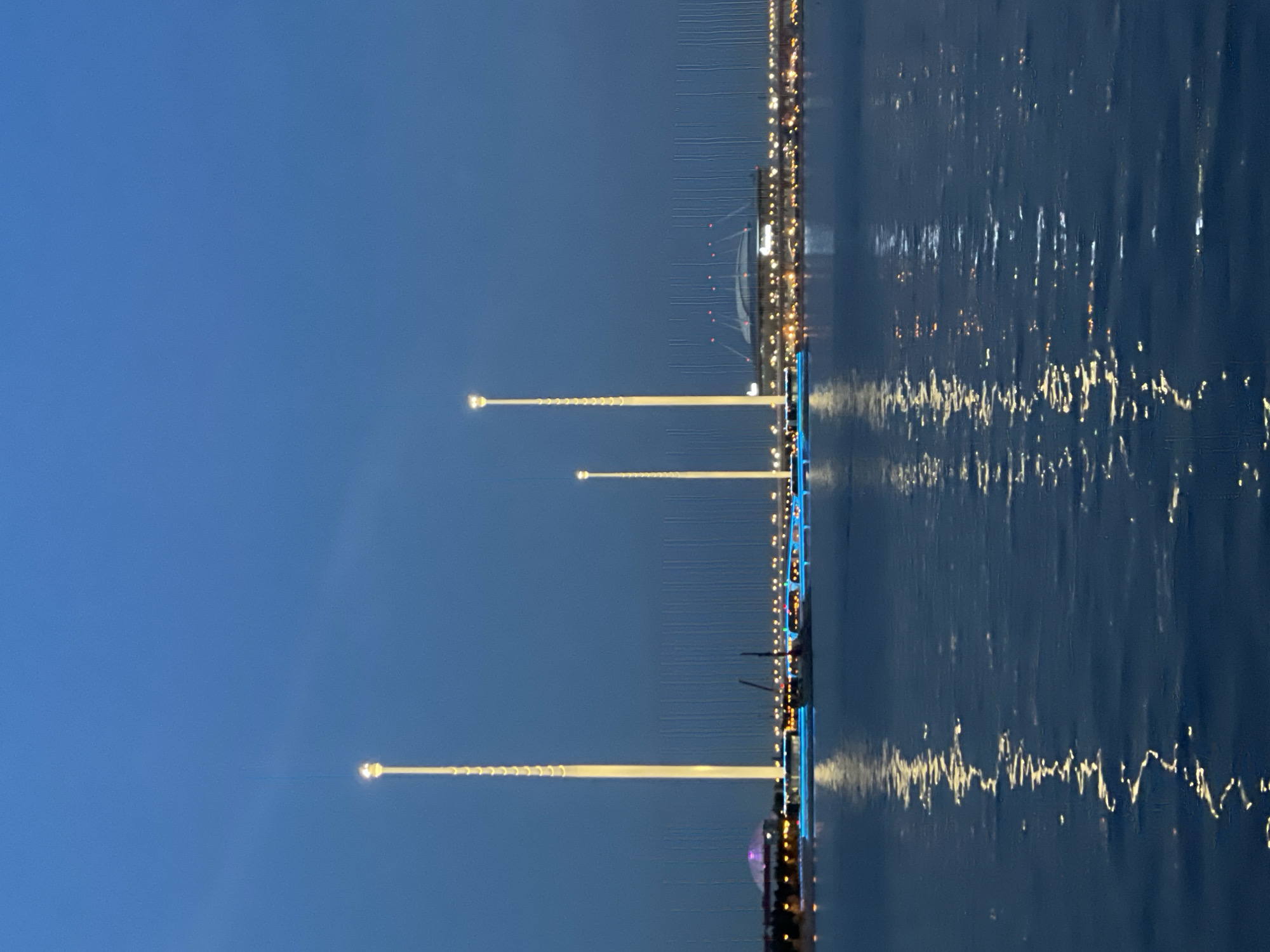 The image depicts a nighttime view of the Gulf of Finland in Saint Petersburg, Russia, with the Krestovsky Stadium and surrounding illuminated structures visible in the distance. Three tall, slender illuminated towers are prominent in the foreground, with the tallest in the center, followed by a slightly shorter one to its left and a shorter one to its right. The towers emit a bright, warm yellow light and appear to be adorned with a spiraling pattern of lights near their tops. The water in the foreground reflects the lights from the towers and the distant cityscape, creating shimmering patterns on its surface.
Across the water, the Krestovsky Stadium is identifiable by its distinctive domed roof and illuminated facade. Adjacent to the stadium, other buildings and infrastructure are lit, forming a luminous line along the coastline. A bridge with blue lighting stretches across the water, connecting parts of the waterfront. A sailboat with its mast visible is positioned in front of the bridge. In the far distance, a rounded dome structure, likely part of another building complex, is also illuminated. The sky is a deep twilight blue, with a few faint vertical lines, possibly indicating some sort of aerial or lighting installation. No people are discernible in the image.