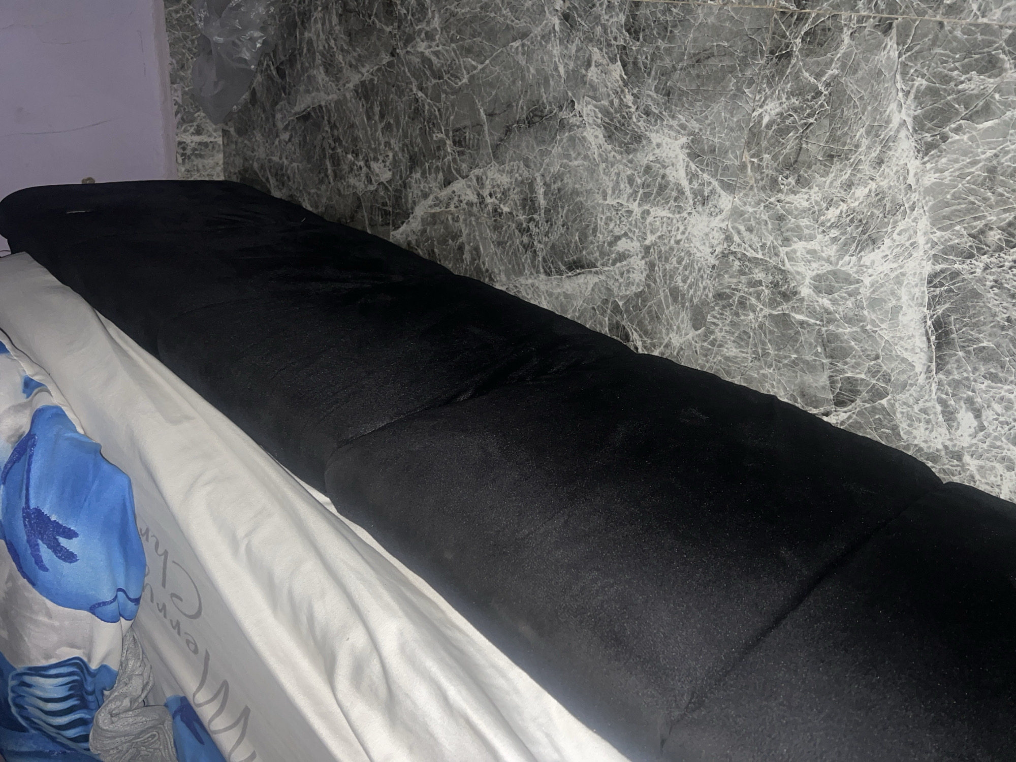 An indoor scene in Jauro Yinu, Nigeria, depicts a bed angled from the top left to the bottom right, positioned against a light purple wall and a grey marble-patterned tiled floor. The bed features a white sheet with faint grey text reading "Merry Chr..." and pillows covered by blue and white patterned cases at its head. The visible side of the bed frame or skirt is black, composed of a textured fabric. The floor is covered with large grey and white veined tiles. A crumpled, clear plastic bag rests on the tiled floor in the upper right quadrant.