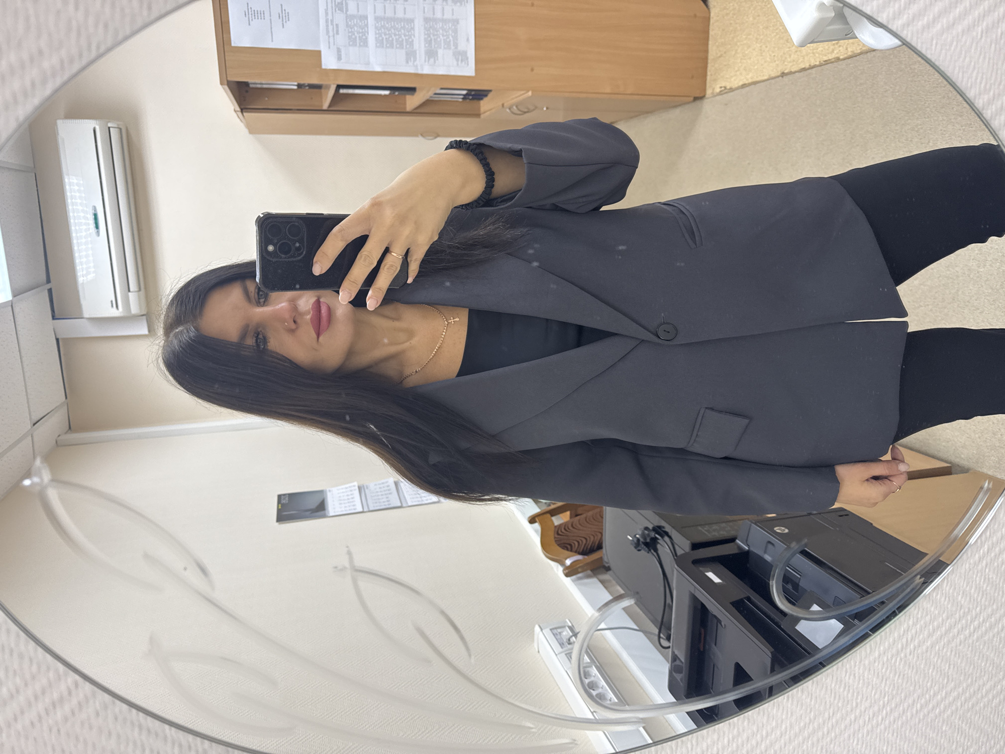 A young to middle-aged woman is taking a mirror selfie in what appears to be an indoor office or workspace setting. The image is captured through a large, round mirror that features frosted decorative patterns on its surface.
The woman has long, dark hair and is wearing a dark, possibly charcoal grey, blazer over a black top, paired with dark pants. A gold chain with a small cross pendant is visible around her neck, and a dark scrunchie or bracelet adorns her right wrist. She is holding a dark smartphone with a prominent camera array, seemingly an iPhone, to capture the image. A ring is visible on her left ring finger. Her expression is neutral, and she is looking directly at the camera. She has light makeup, including pink lipstick, and a small nose piercing.
The background reflected in the mirror shows a light-colored wall with an air conditioner unit mounted on it to the left. To the right, a wooden shelf or cabinet is visible, with several sheets of paper containing typed text, possibly schedules or documents, tacked onto it. On a lower surface, a black printer is discernible, next to a white power strip with multiple outlets and various cables connected. The floor appears to be carpeted in a light, neutral color. The overall lighting suggests daytime. The text on the papers in the background is not legible.
