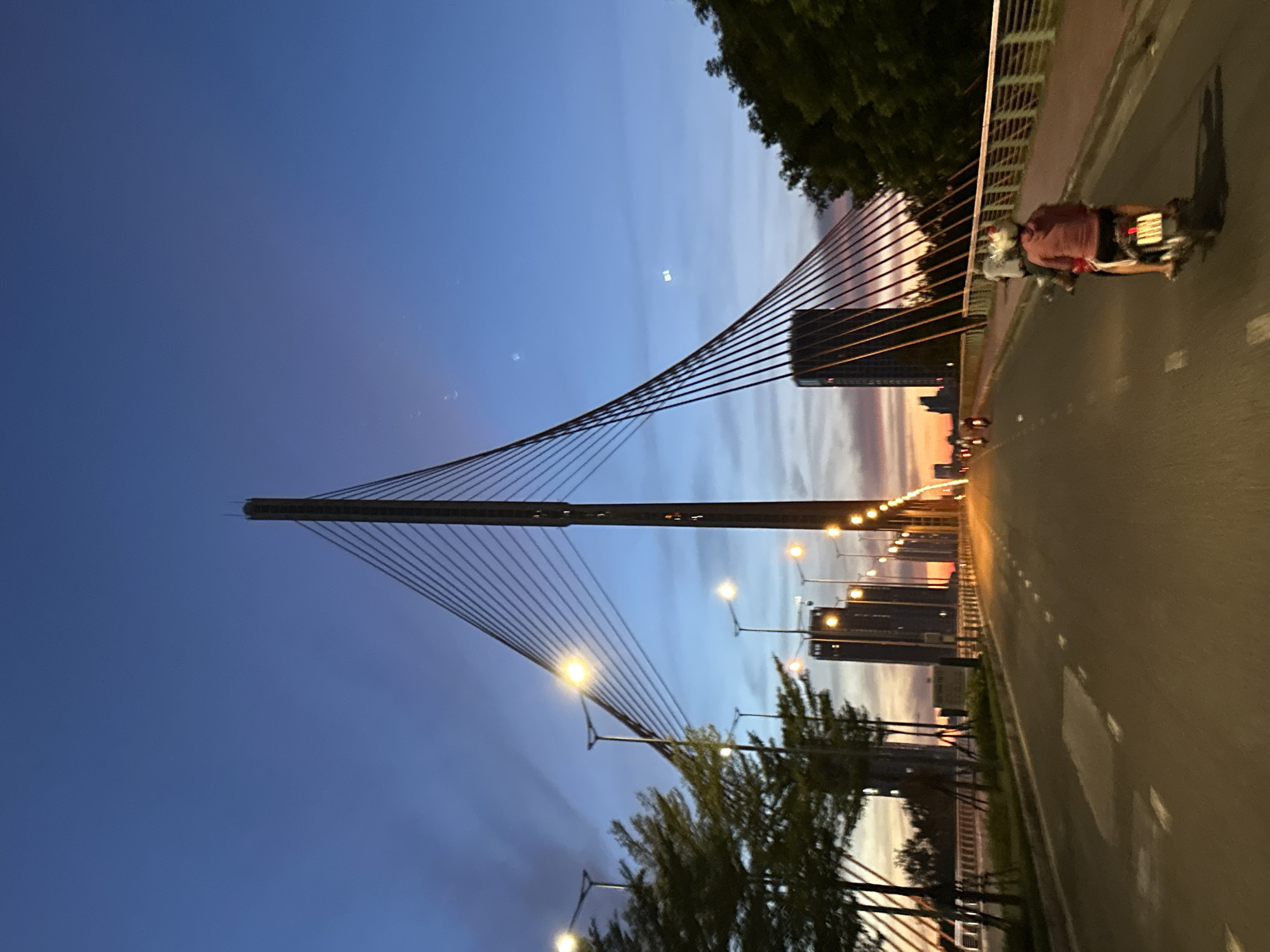 The image captures a sunset view of the Dragon Bridge in Da Nang, Vietnam, at dusk. A person is riding a motorcycle in the foreground, with another person seated behind them, both wearing helmets. The motorcycle is traveling on the bridge's roadway, which is illuminated by streetlights and marked with white dashed lines. Tall buildings and trees line the sides of the bridge, and the sky displays a gradient of blue and orange hues, indicating the setting sun. The bridge's iconic structure, with its tall central pylon and suspension cables, is a prominent feature in the background. The scene is calm, with a few other motorcycles visible in the distance.