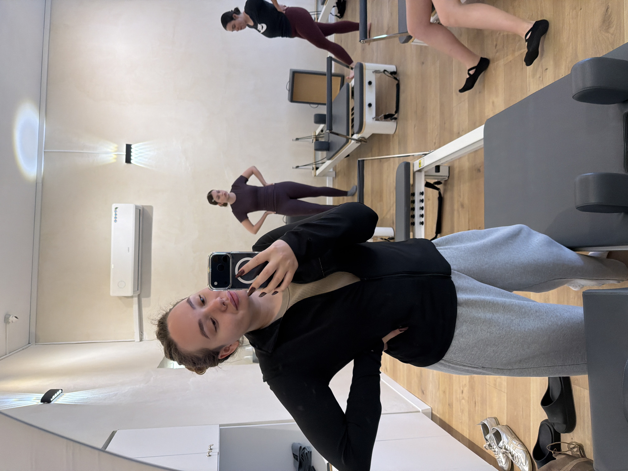 The image is taken in a mirrored Pilates studio in Ramat Gan, Israel. A woman takes a selfie in a mirror, wearing grey sweatpants and a black jacket. Reflected in the mirror are two other women practicing Pilates on reformer machines. One woman wears a black top and burgundy leggings, and the other wears a purple outfit. Gym shoes and casual shoes are on the floor. There is an air conditioner and modern light fixture on the wall.