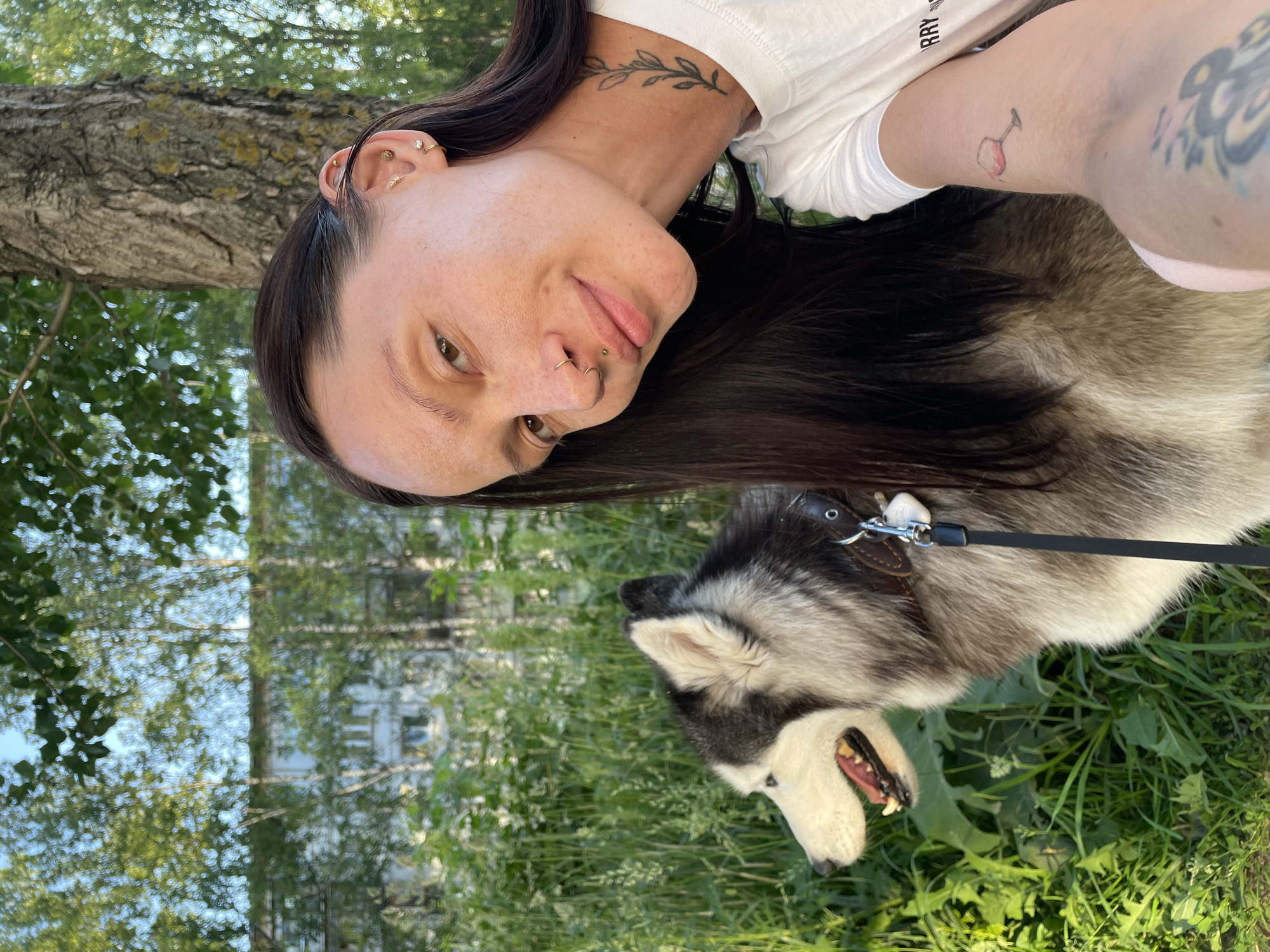 The image shows a woman and her dog, a husky, outdoors in a grassy area with trees and a building in the background. The woman is taking a selfie, with her face close to the camera. She has dark hair, visible tattoos on her neck and arm, and piercings on her ear and nose. The husky is panting with its mouth open, looking to the side. The background suggests a park or residential area with greenery and a building that appears to be an apartment block. The lighting indicates it is daytime, likely sunny. The woman's expression is neutral, and the dog appears relaxed. The text on the woman's sleeve is partially visible and says "SORRY". The scene is set in Izhevsk, Russia.