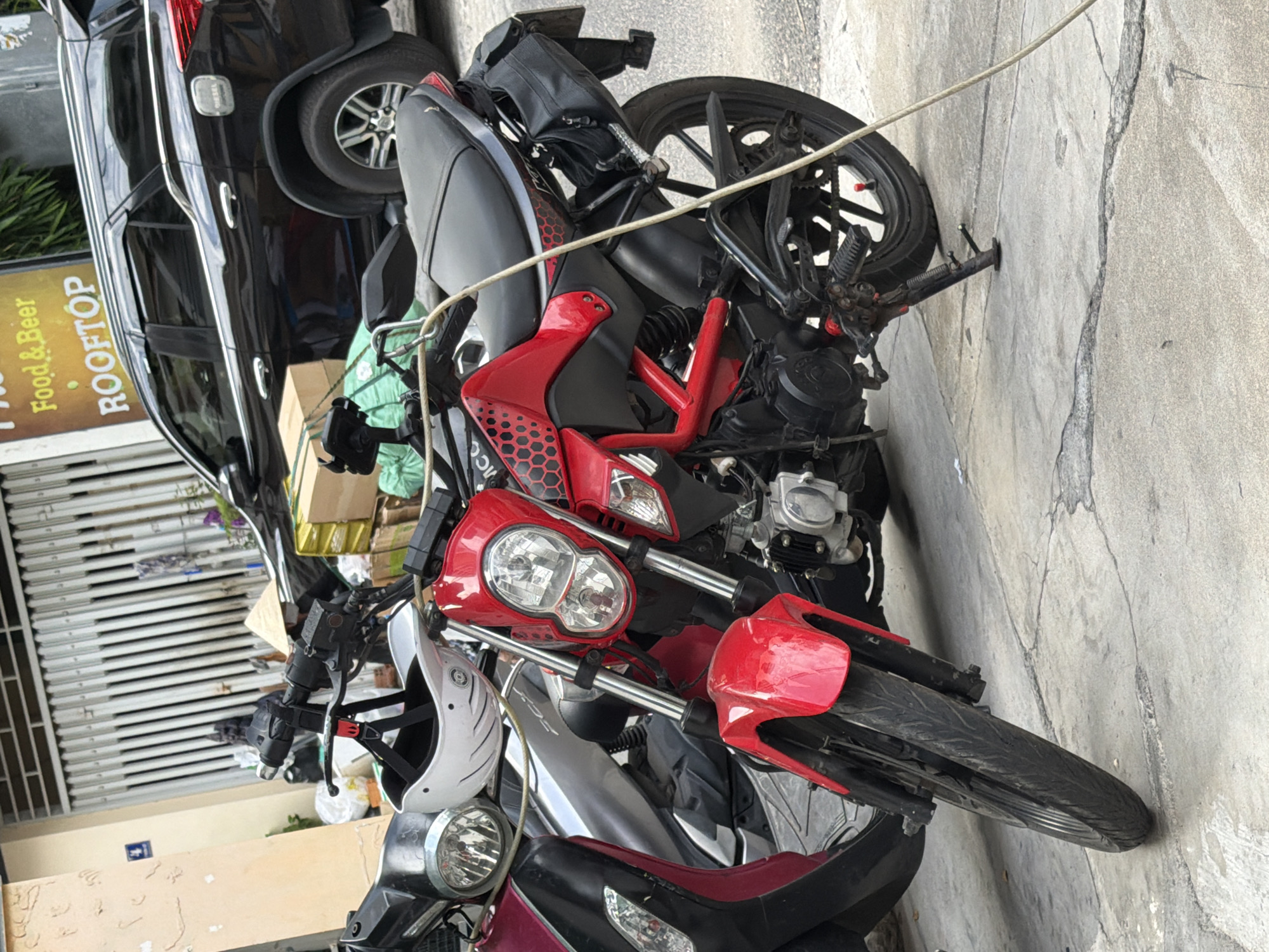 The image depicts a street scene in Nha Trang, Vietnam, likely in a commercial or residential area. In the foreground, a red and black motorcycle is parked on a concrete surface. The motorcycle has a distinctive design with a prominent red frame and black accents, including a hexagonal pattern on the bodywork. A white helmet is resting on the handlebars. To the left of the red motorcycle, another scooter, partially visible, is parked. In the background, a black SUV is parked next to a building with a corrugated metal fence. A yellow sign with text that appears to read "Food & Beer ROOFTOP" is visible on the building. There are stacks of boxes and bags of merchandise near the vehicles and building, suggesting commercial activity or goods being stored or transported. The environment appears to be outdoors, with a street and sidewalk visible. The overall impression is a typical urban setting in Vietnam, with motorcycles being a common mode of transportation and businesses operating along the street.