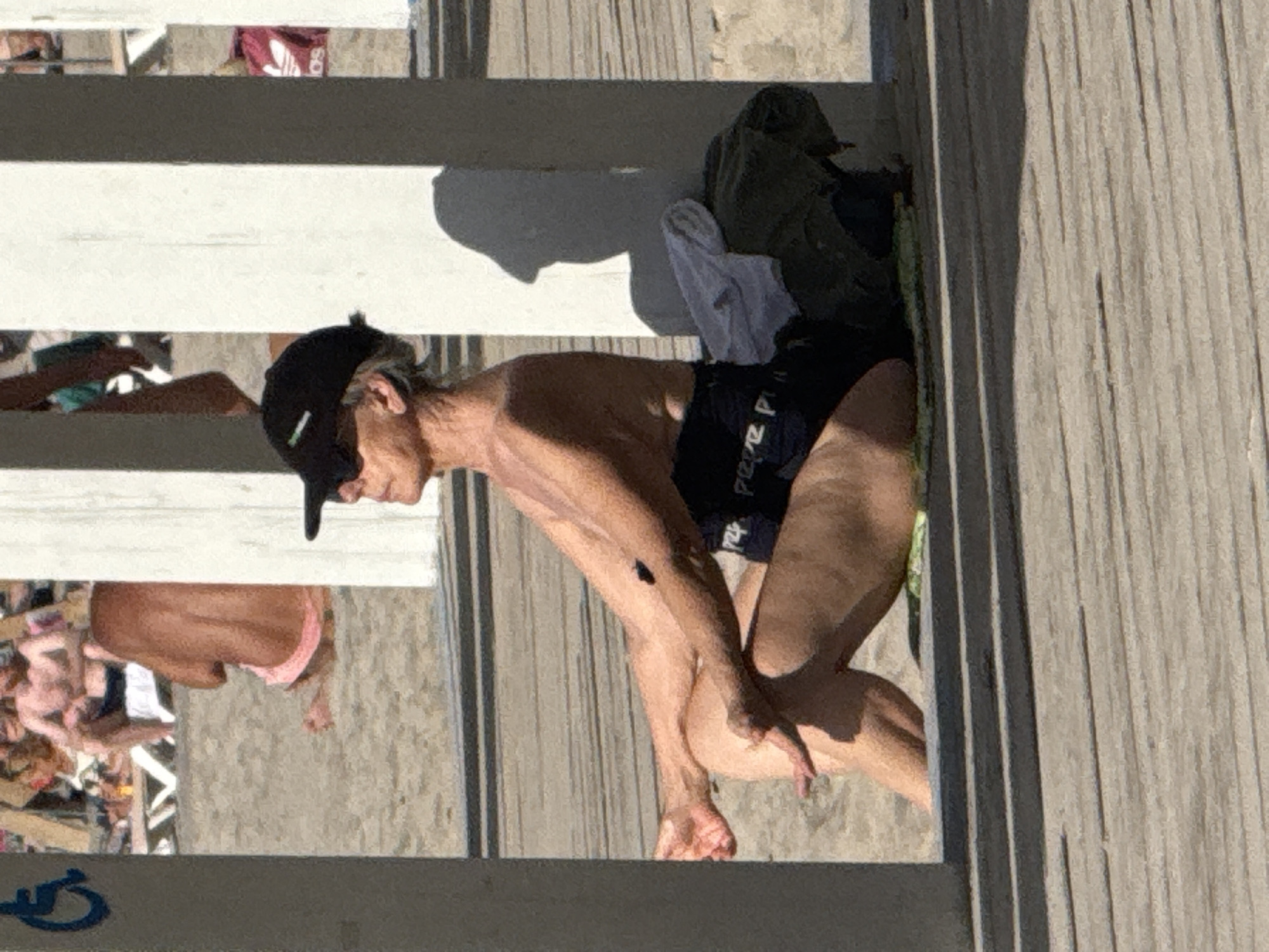 A woman in a black baseball cap and black bathing suit sits on a wooden boardwalk with her legs crossed, facing right. Her left arm is bent and resting on her knee, while her right arm is extended and resting on the ground. Behind her, a pile of clothes is visible, including a white towel and a green garment. In the background, several other people are visible on a sandy beach. The scene appears to be set in a city, though the specific location is unknown. The overall lighting suggests a sunny day.