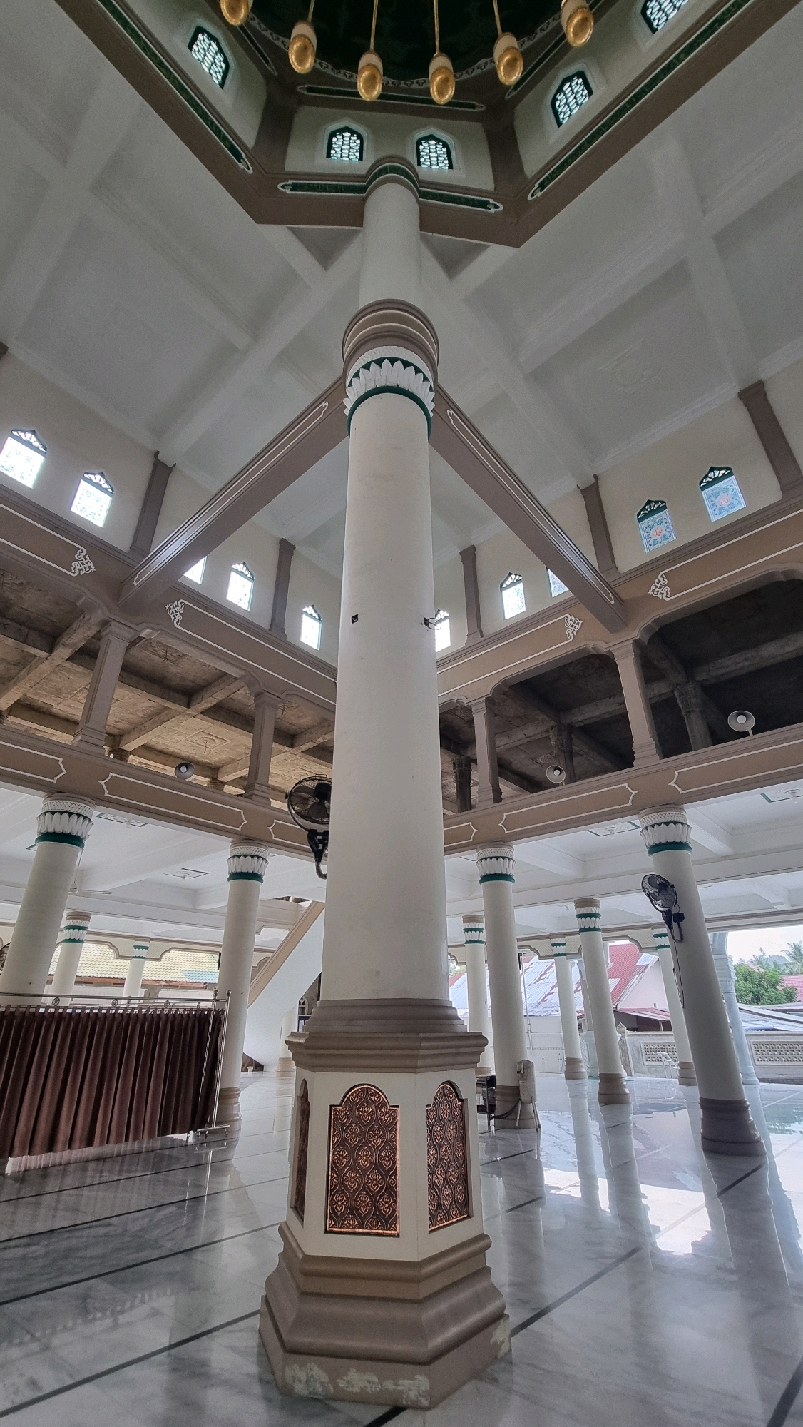 The image shows the interior of a mosque in Lhokseumawe, Indonesia, with a wide-angle perspective emphasizing the central pillar and the surrounding architecture. The mosque features a high, vaulted ceiling with decorative elements, including golden light fixtures and arched windows with intricate latticework. Several large, cream-colored pillars support the structure, some with decorative bands of green and white near their capitals. The floor is made of polished marble with a reflective surface. In the background, brown curtains partially obscure an area, and further back, additional pillars and open archways reveal more of the mosque's interior structure, including exposed concrete beams and ceiling. Fans are mounted on some of the pillars. The overall impression is one of a spacious and well-decorated religious space.