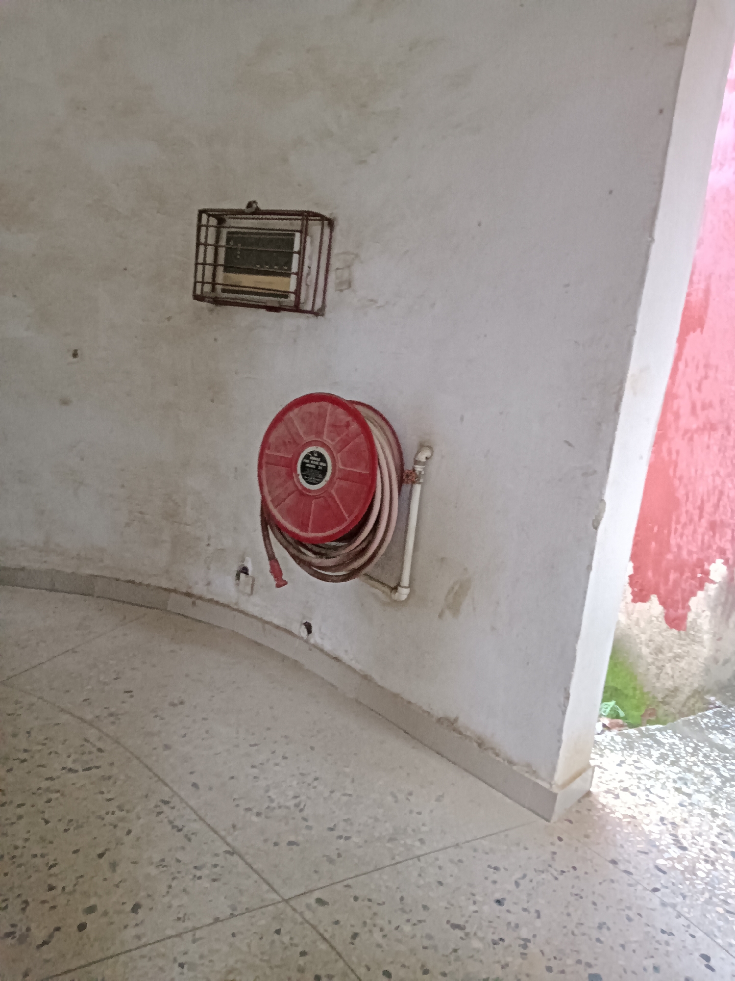 The image depicts an indoor scene with a curved wall and a tiled floor. Mounted on the wall are two objects: a metal grate-covered item with visible internal components, possibly a heater or ventilation unit, and a red fire hose reel with a coiled hose and nozzle. The wall appears weathered, with some discoloration and texture. The floor is made of terrazzo or a similar composite material, with visible grout lines forming a pattern. To the right of the wall, a brightly colored pink surface is partially visible, suggesting an adjacent space or exterior wall. Sunlight casts shadows on the floor, indicating a light source from the right. No people or animals are present in the scene.