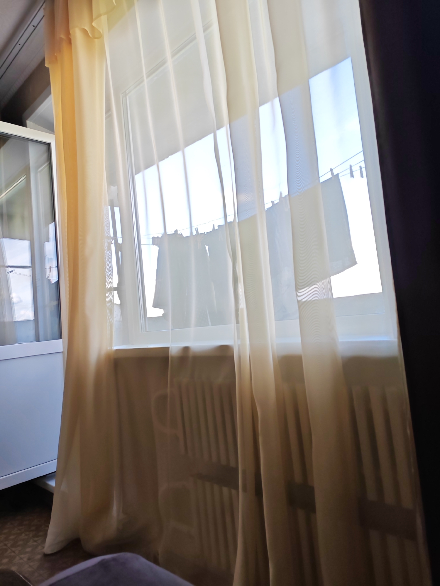 A photograph taken indoors, likely in a residential setting in Smolensk, Russia, captures a view through a window covered by sheer, cream-colored curtains. The curtains are illuminated by bright sunlight streaming from outside, creating a hazy, diffused light within the room. Through the translucent fabric, clothing items are visible hanging on a clothesline outside the window, suggesting laundry is being dried. A radiator is positioned beneath the window, its vertical slats visible through the curtain. To the left of the window, a portion of a white cabinet with a glass door is present. The floor appears to be tiled. A dark, opaque curtain is visible on the right side of the frame. The overall impression is one of a domestic scene on a sunny day, with a focus on the interplay of light and fabric.