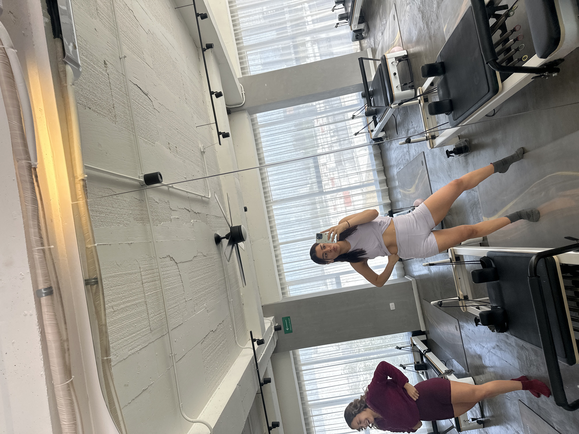 The image depicts two women in a brightly lit room, likely a Pilates studio, given the presence of multiple Pilates reformers. The studio appears to be located in Puebla, Mexico, suggested by the context of the original prompt.
The woman on the right, closer to the viewer, is taking a selfie in a large mirror that covers a significant portion of the wall. She is wearing a light purple athletic set, consisting of a crop top and shorts. Her left hand is on her hip, and her right hand holds a smartphone, angled to capture her reflection. She has dark hair pulled back and is looking at her phone.
The second woman is positioned to the left of the first, also in the reflection of the mirror. She is wearing a dark red or maroon athletic set, including a long-sleeved top and shorts. She is looking to her left and appears to be laughing or smiling.
The room features large windows with sheer white curtains, allowing natural light to enter. Several Pilates reformers are arranged in rows. The ceiling is white and exposed in some areas, with visible pipes and lighting fixtures, including a ceiling fan. The floor is a polished concrete. A green exit sign is visible on a grey wall in the background. The overall atmosphere is clean and functional, typical of a fitness facility.