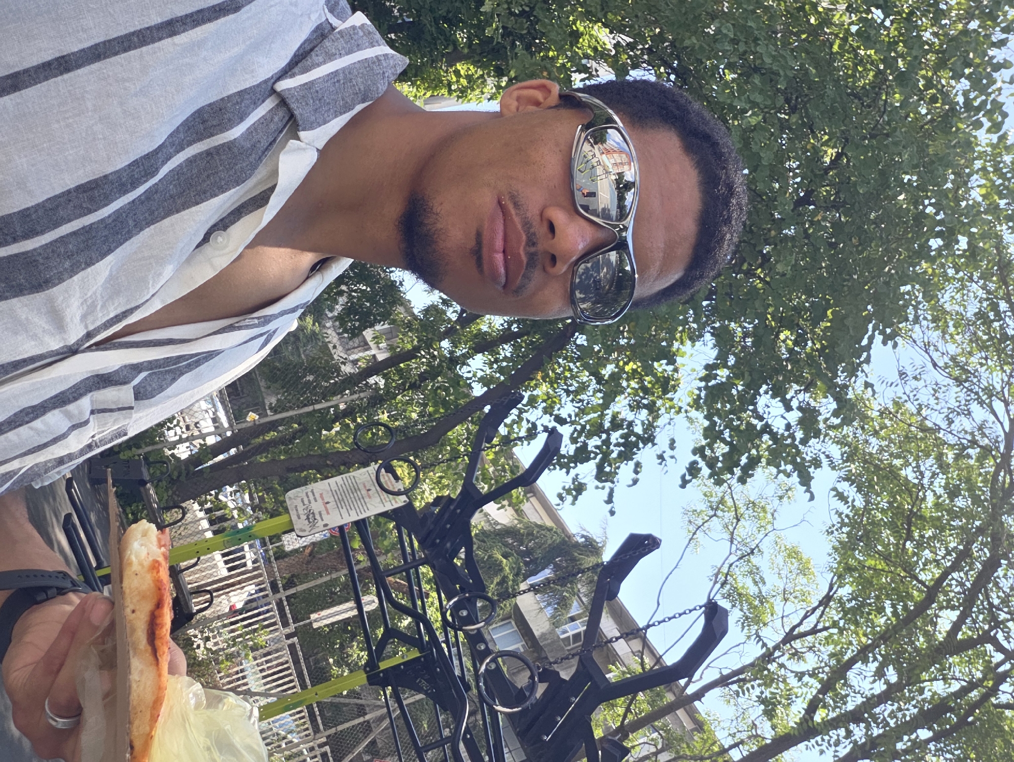 A young Black man with short, curly hair and a goatee is taking a selfie outdoors. He is wearing silver-rimmed sunglasses and a white and gray striped button-down shirt. In his right hand, he holds a slice of pizza, wrapped in a light yellow plastic bag and resting on a cardboard base.
The background reveals a vibrant urban setting, characterized by lush green trees and a clear blue sky. To the right, a black metal structure with rings suggests a playground or outdoor fitness equipment. In the distance, buildings with windows are visible, hinting at a residential area. The reflection in the man's sunglasses shows a street scene with buildings and a traffic light.
The man's gaze is directed towards the camera, and his expression is neutral. The overall scene depicts a casual moment outdoors, possibly in a park or public space within the city of Varna, Bulgaria. The presence of trees and the style of architecture in the background might further indicate a specific neighborhood or area within Varna.