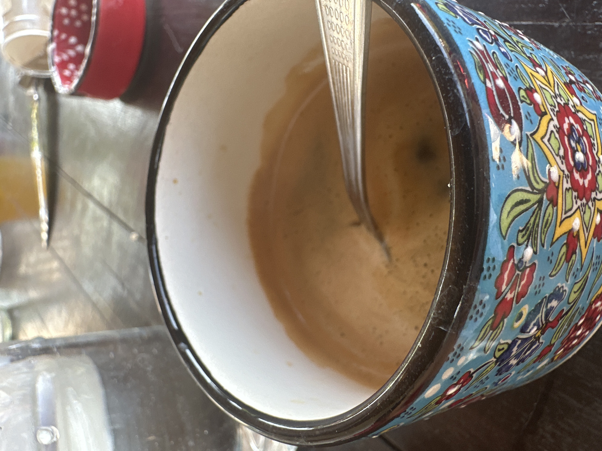 A close-up shot captures a vibrantly decorated mug filled with coffee, resting on a dark, textured surface. A silver spoon is partially submerged in the coffee. In the background, a glass of water and a small red dish with white polka dots are visible, suggesting a cafe or restaurant setting. The mug itself is adorned with intricate blue, red, yellow, and green floral patterns. The scene does not provide any specific visual cues to indicate its location in the city of Galiche, Bulgaria.