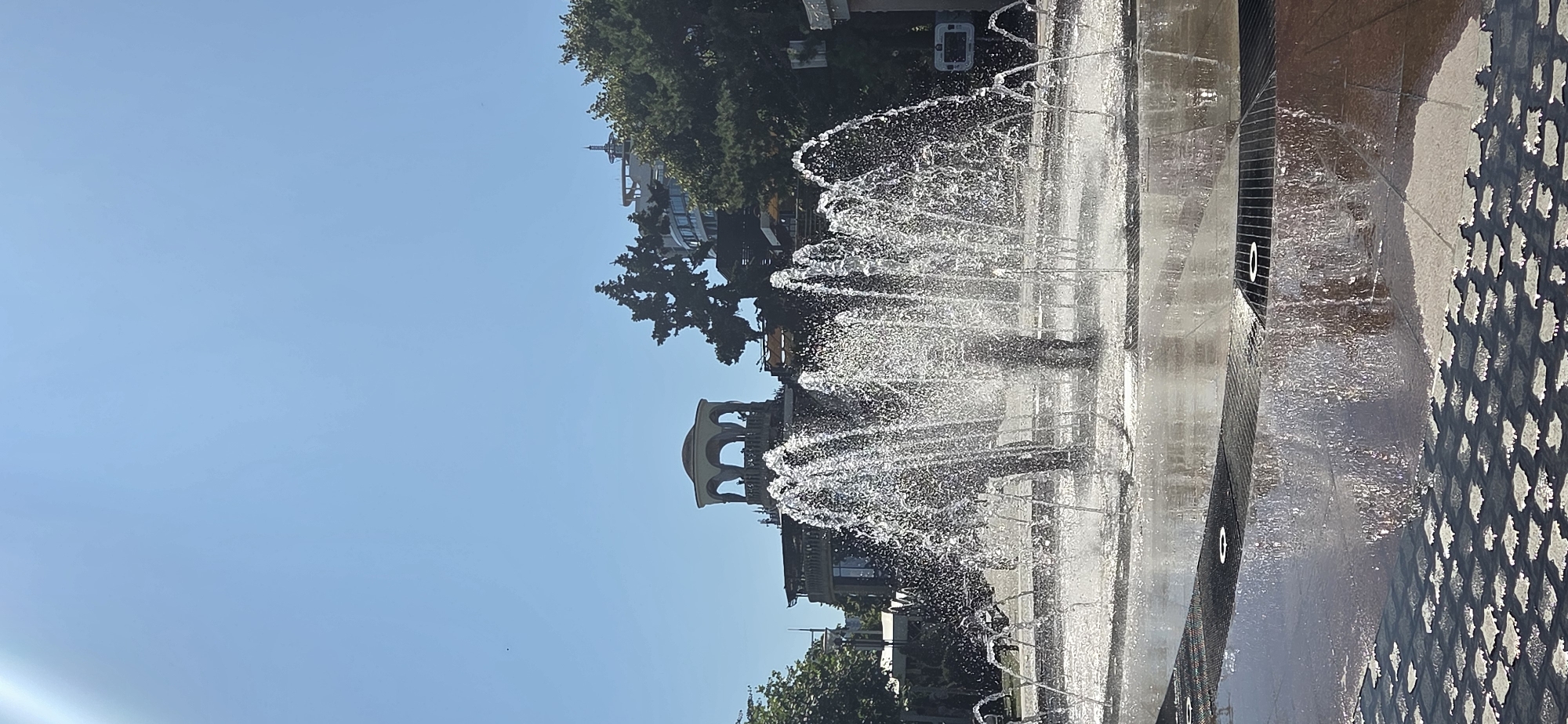The image depicts a sunny day in Rostov-on-Don, Russia, with a focus on a large, active fountain in the foreground. The fountain is the central subject, with multiple streams of water spraying upwards and outwards, creating a dynamic display. The water reflects the sunlight, creating a shimmering effect.
In the background, various architectural elements are visible. To the left, there is a building with a distinctive rotunda-like structure, featuring arched openings and a domed roof. To the right of the rotunda, a tall, dense evergreen tree stands prominently. Further to the right, another building with a modern, curved facade and a circular element on its roof is visible, partially obscured by foliage. There are also hints of other buildings and trees, suggesting an urban park or public square setting.
The ground surrounding the fountain appears to be paved with dark tiles, with some sections featuring a patterned texture and circular grates. The wet surfaces of the fountain base and surrounding pavement indicate the presence of water. The sky above is a clear, bright blue, signifying good weather.
A few individuals are faintly visible within the spray of the fountain, appearing as dark silhouettes, suggesting they are standing in or near the water. The overall scene captures a lively public space on a clear, warm day.