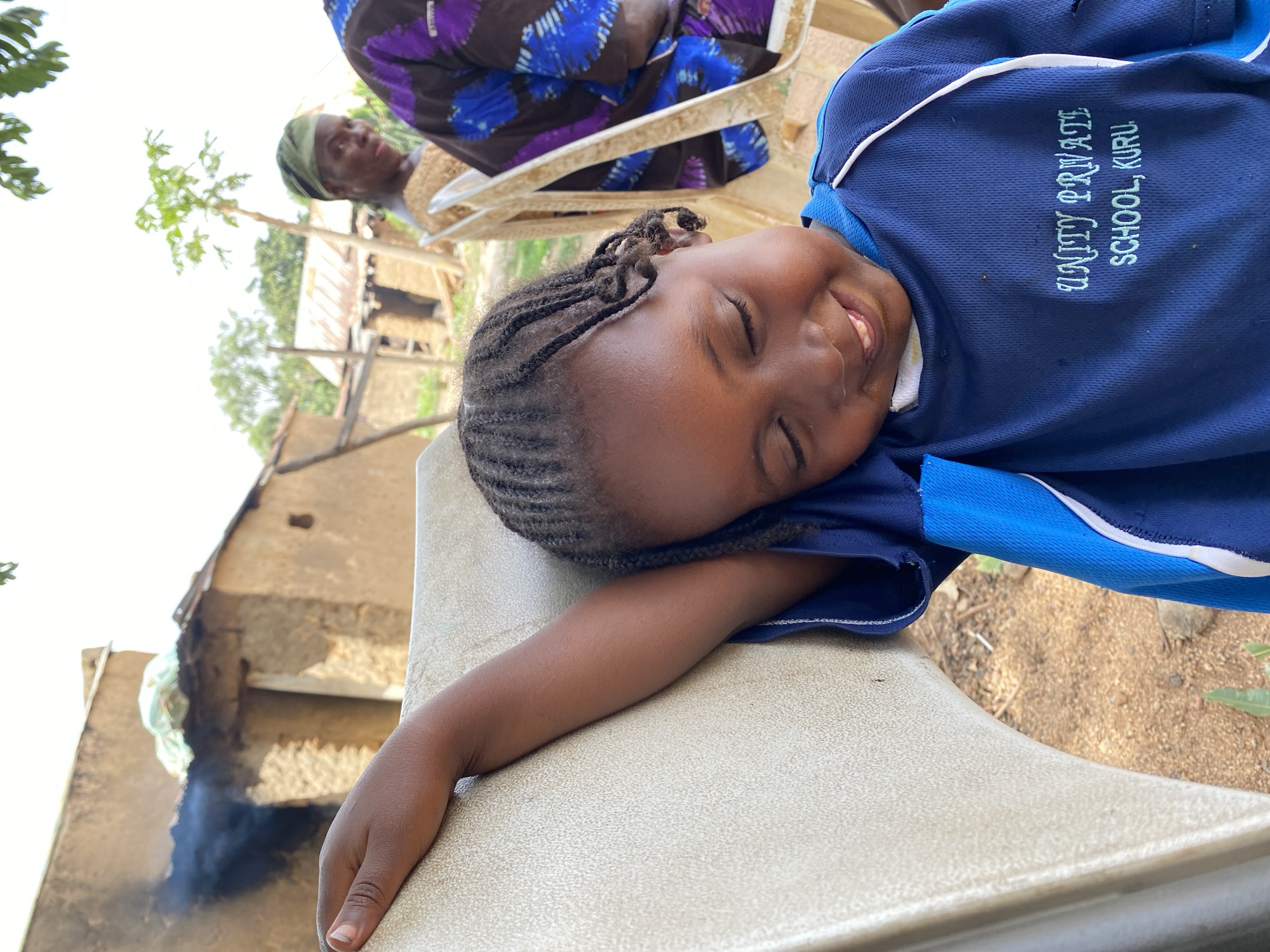 A young girl wearing a navy blue shirt with “UNITY PRIVATE SCHOOL, KURU” printed on the front is lying on a beige surface, possibly a chair or table, with her eyes closed and a slight smile on her face. Her dark hair is styled in braids. Behind her, an older woman with a green head covering is sitting in a chair, partially visible in the upper portion of the frame. In the background, there are glimpses of what appears to be a rural setting, with a weathered building, vegetation, and smoke in the air. The location is indicated by the school name, suggesting the scene is in Kuru Dungus, Nigeria.
