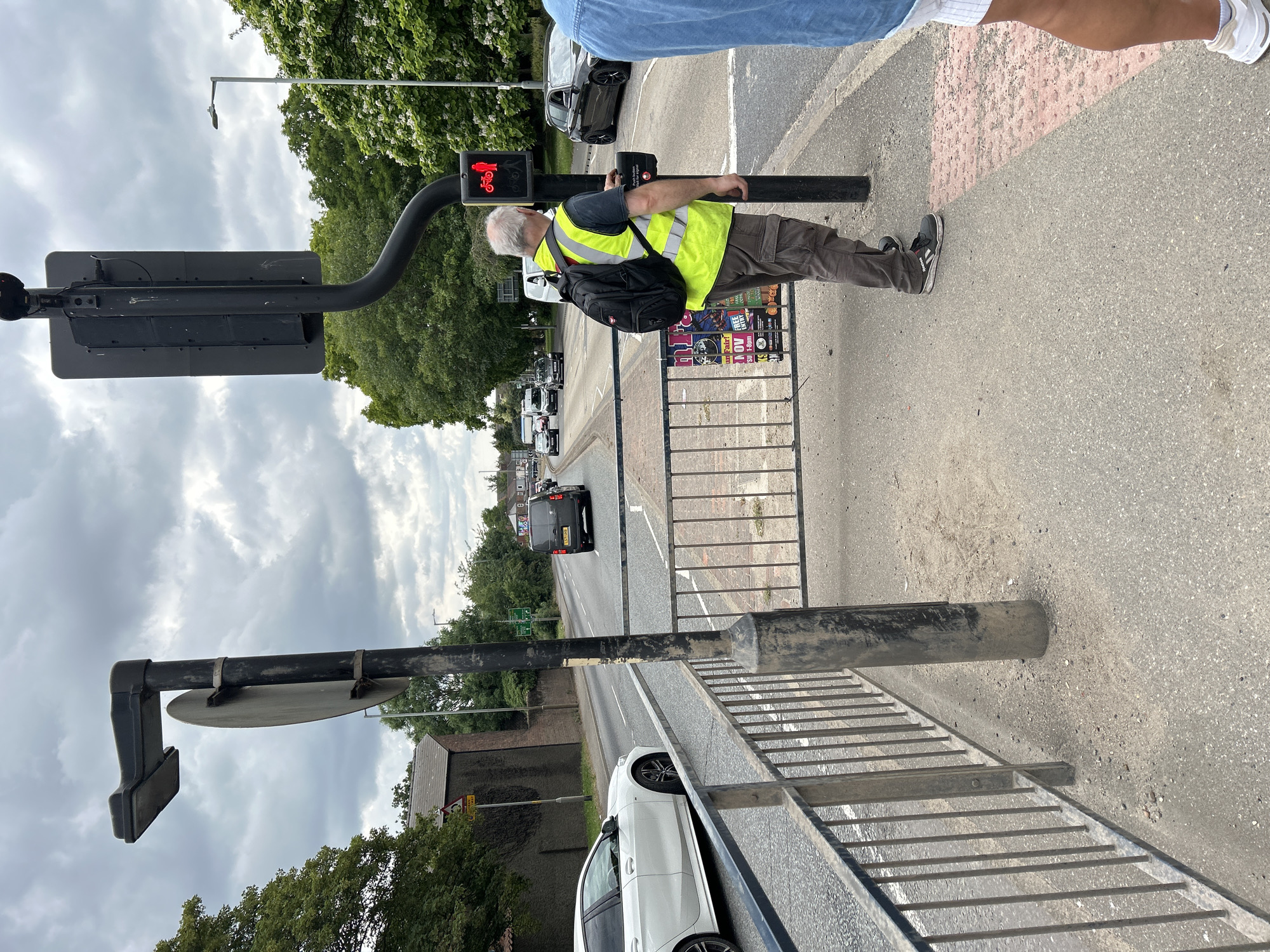 The image depicts a street scene in Boston, United Kingdom, with a focus on a pedestrian crossing. A man wearing a high-visibility yellow vest and a backpack stands at a traffic light, facing the street. He appears to be waiting to cross, as indicated by his proximity to the crossing signal which displays a red cyclist symbol. The traffic light pole is black and curves from a vertical position to horizontally over the crossing. A push-button unit is attached to the pole at a height accessible to pedestrians.
To the left of the man, a metal barrier fence runs along the pavement. Beyond the fence is a road with several cars driving on it. A white car is visible on the left side of the road. In the background, trees line the street, and buildings are visible further down. The sky is cloudy, suggesting overcast weather.
The man's attire, the style of the traffic lights, and the presence of a push-button for the crossing are consistent with urban infrastructure found in the United Kingdom. The overall environment suggests a typical suburban or urban street.