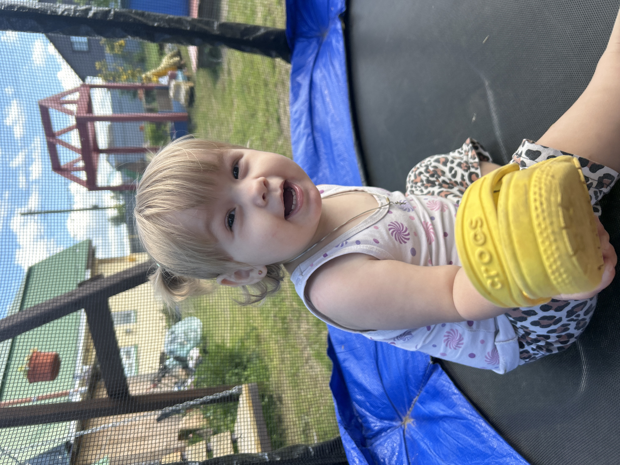 A young blonde girl with pigtails sits on a trampoline, holding a yellow Croc shoe. She is wearing a white tank top with a pink swirled pattern and leopard print shorts. The background shows a backyard with houses, a red play structure, and a swing set, under a blue sky with clouds. The location is identified as Olkhovka, Russia.