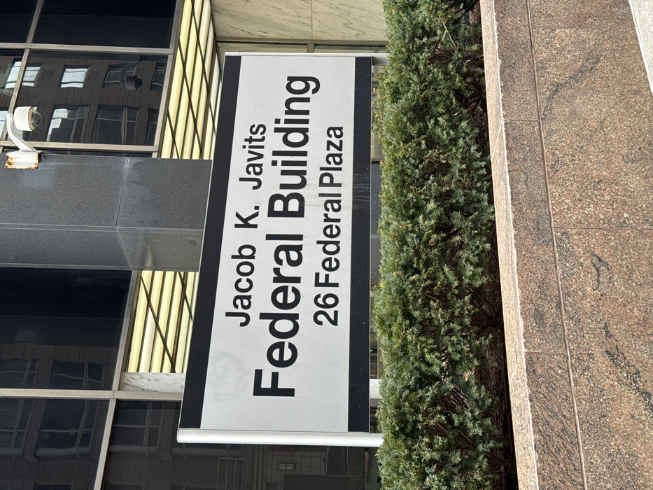 A sign for the Jacob K. Javits Federal Building at 26 Federal Plaza is affixed to the building's exterior. The sign has white text on a grey background and is oriented vertically. Next to the sign is a narrow evergreen tree. The building's facade is composed of a combination of dark grey stone, light tan stone and glass. A security camera is mounted to the building.