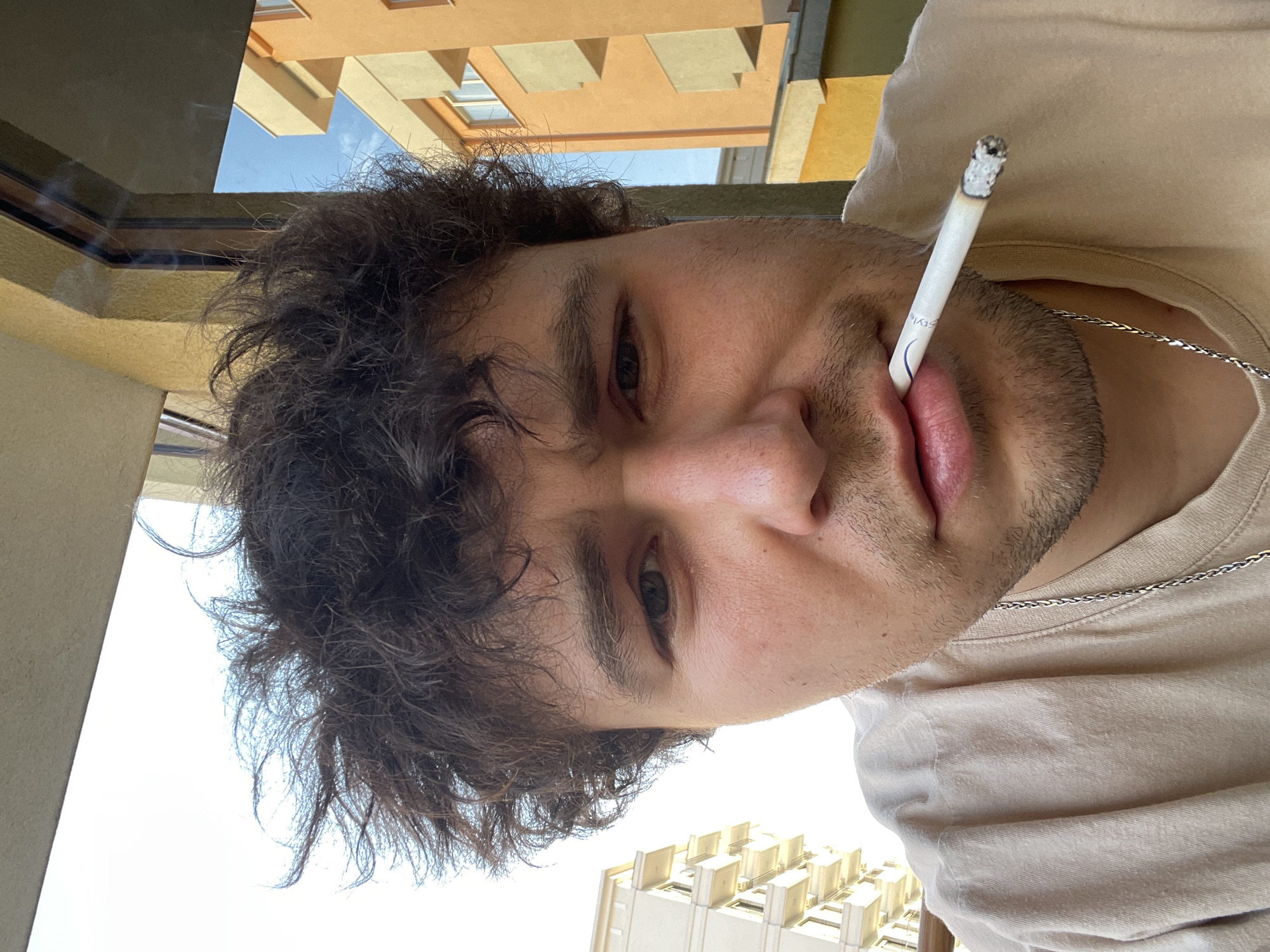 A head-and-shoulders view of a fair-skinned male with dark, curly hair is shown holding a lit cigarette in his mouth. He is wearing a beige T-shirt and a silver necklace. Buildings are visible in the background, suggesting an urban environment. The image may have been taken in Bar, Montenegro.