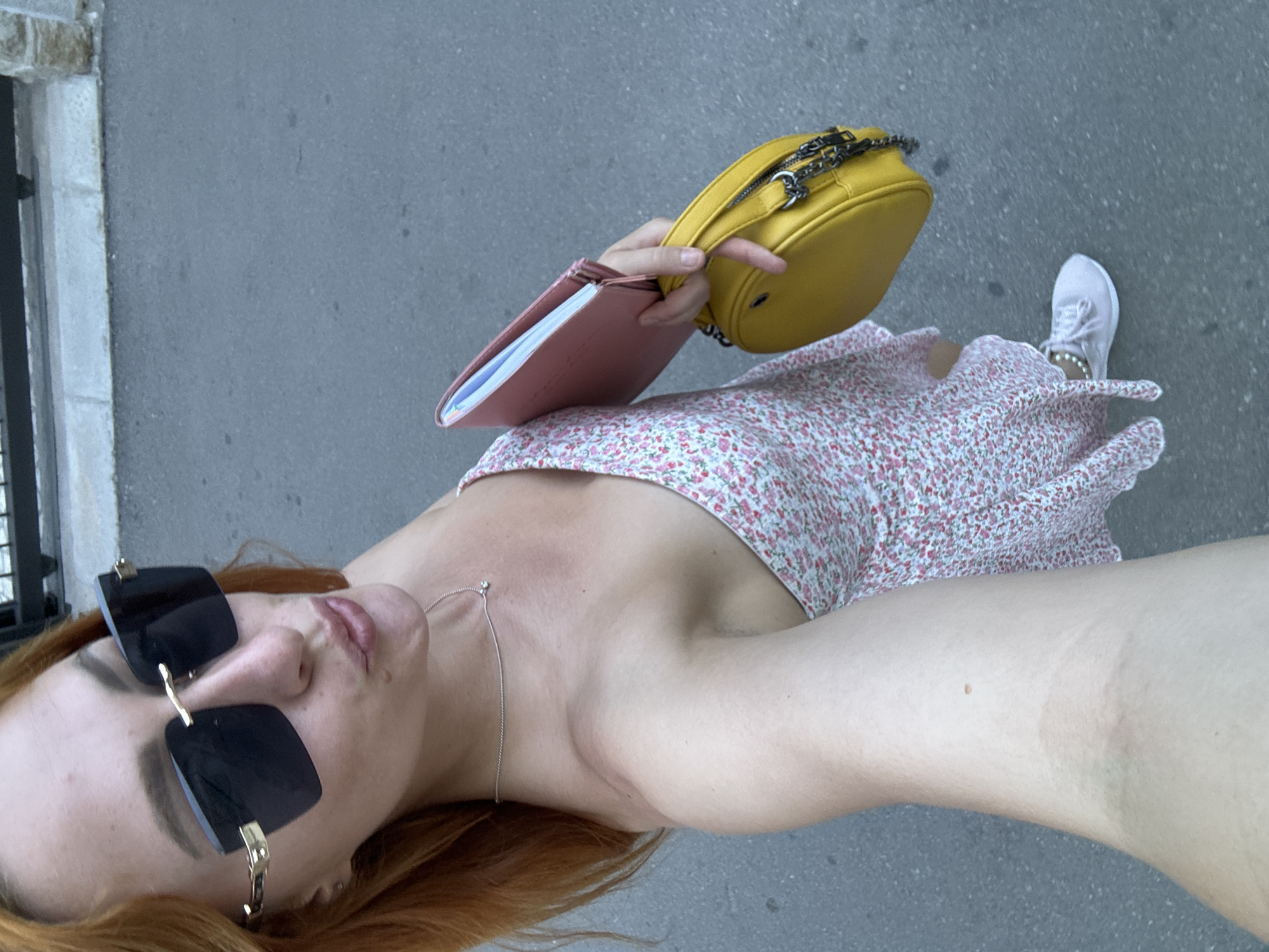 In Budva, Montenegro, a young woman with reddish-brown hair is captured in a selfie from a high-angle perspective. She is wearing large, square, black-rimmed sunglasses with gold accents and a delicate silver chain necklace with a small pendant. Her lips are slightly pursed. She is dressed in a floral print dress with a light background and small pink and red flowers. She is carrying a pink notebook or binder in her left hand, and a yellow, rounded handbag with a silver chain strap in her right hand. She is wearing light pink sneakers. The background consists of a paved surface, likely a sidewalk or road, and a low stone wall with a metal railing above it, suggesting an outdoor urban environment. The lighting indicates it is daytime.