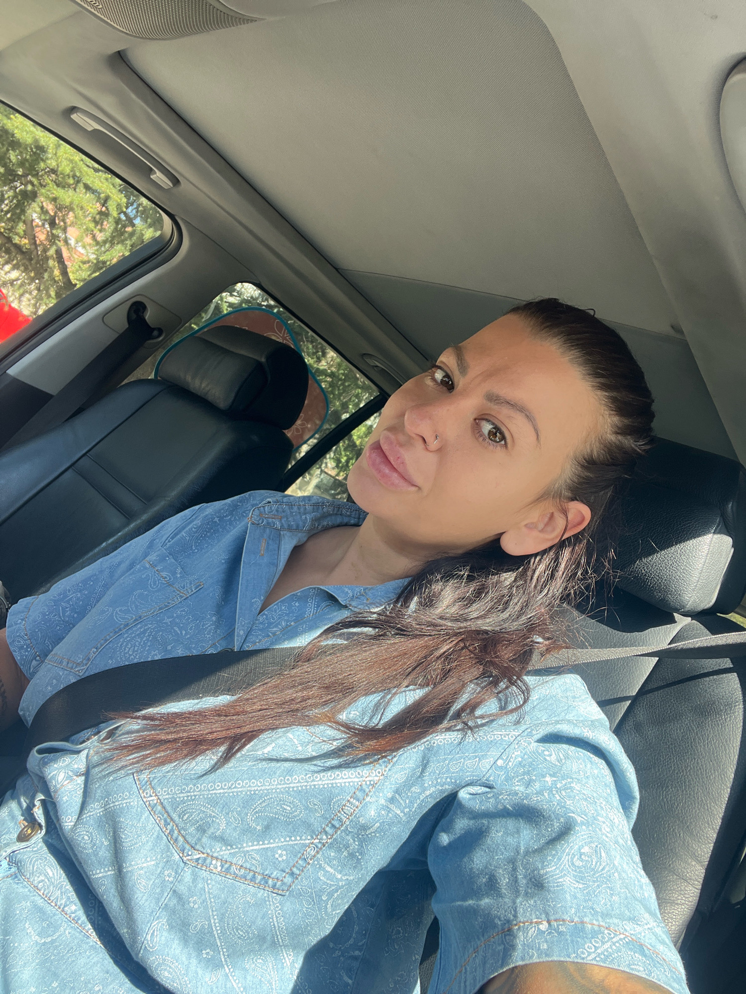 A woman with dark hair pulled back is sitting in the passenger seat of a car, looking towards the camera. She is wearing a light blue denim shirt with a paisley pattern. Her seatbelt is fastened across her chest. The interior of the car is visible, including black leather seats and the headrest behind her. Sunlight streams in through the window, casting shadows on her face and clothes. The background shows trees and foliage, suggesting an outdoor environment, possibly on a street in Plovdiv, Bulgaria, though no specific landmarks are identifiable.