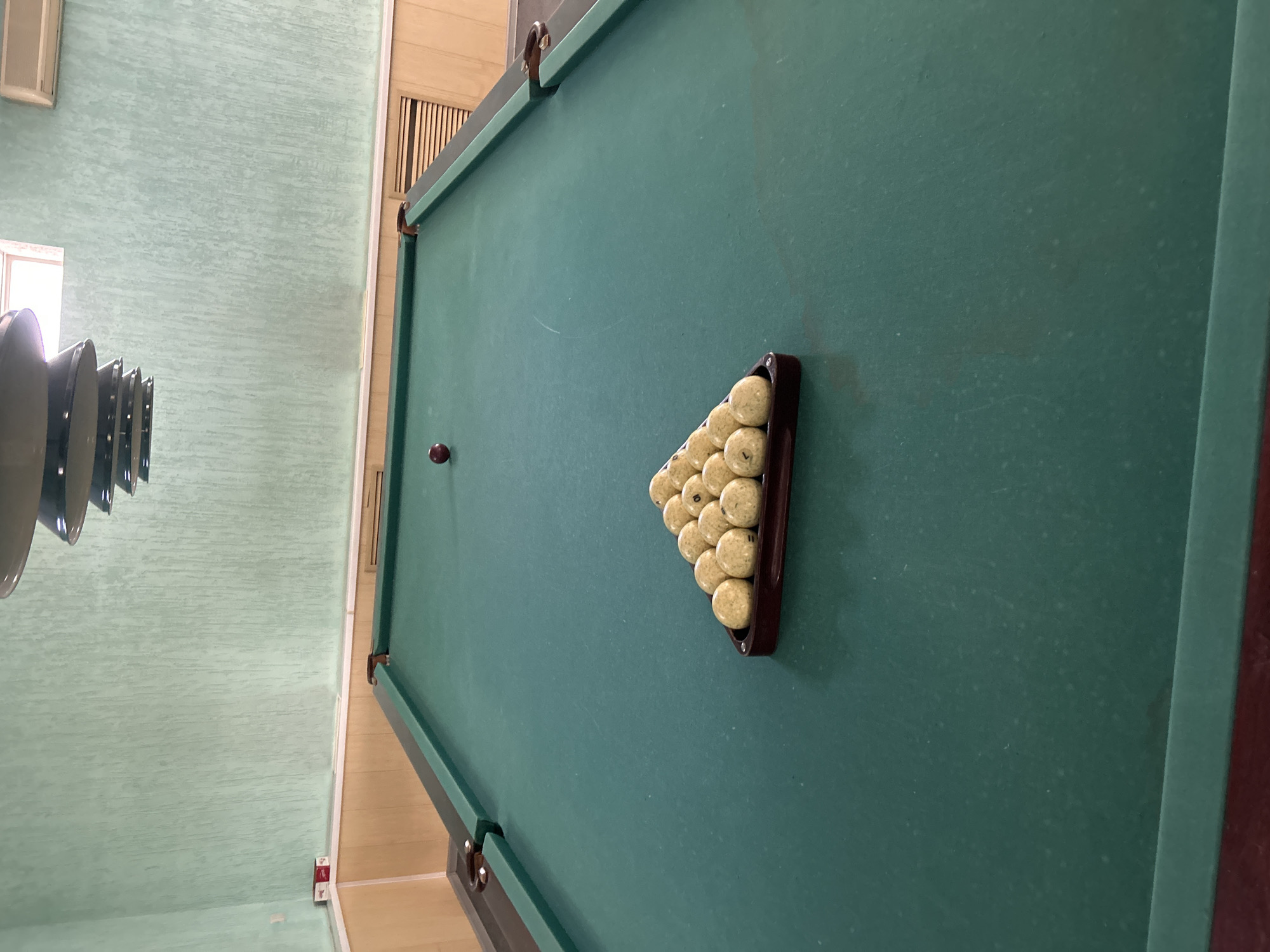 The image shows a billiard table with a rack of billiard balls arranged in a triangle. The table is green, and the balls are light-colored with some numbers visible on them. There is also a single red billiard ball on the table. The table appears to be in a room with light green walls and wooden paneling near the floor. A portion of a ceiling fixture is visible at the top of the image, and an air conditioning unit is mounted on the wall. The image was taken in Chaikovsky, Russia.