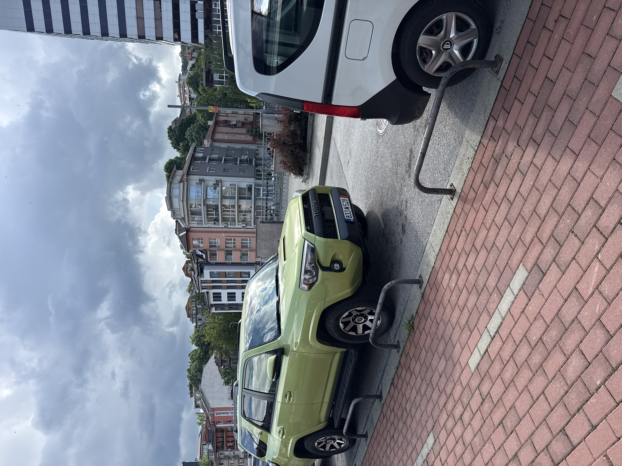 The image shows a lime-green Toyota 4Runner parked on a street in Gabrovo, Bulgaria. The vehicle's license plate reads "833 M528." It is parked next to a white van. The street is paved with red bricks and lined with metal bollards. Buildings of varying heights and architectural styles are visible in the background. The sky is cloudy.