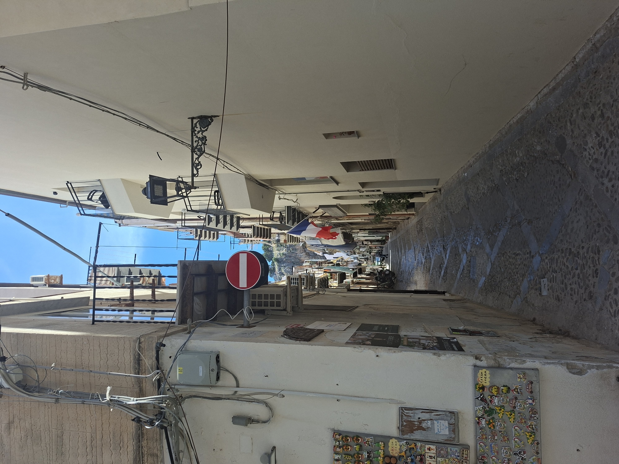 Here's a description of the image:
The image is taken from within a narrow alleyway in Cefalù, Italy. The alleyway is flanked by buildings with white walls and various attachments like electrical boxes, air conditioning units, and a "do not enter" sign. Souvenirs and advertisements are affixed to the walls. Overhead, there's a network of electrical wires and various architectural features. In the distance, the alleyway opens up, revealing a glimpse of a French flag and the harbor. The alleyway is paved with cobblestones. The image suggests a sunny day. There are no people visible within the alleyway.