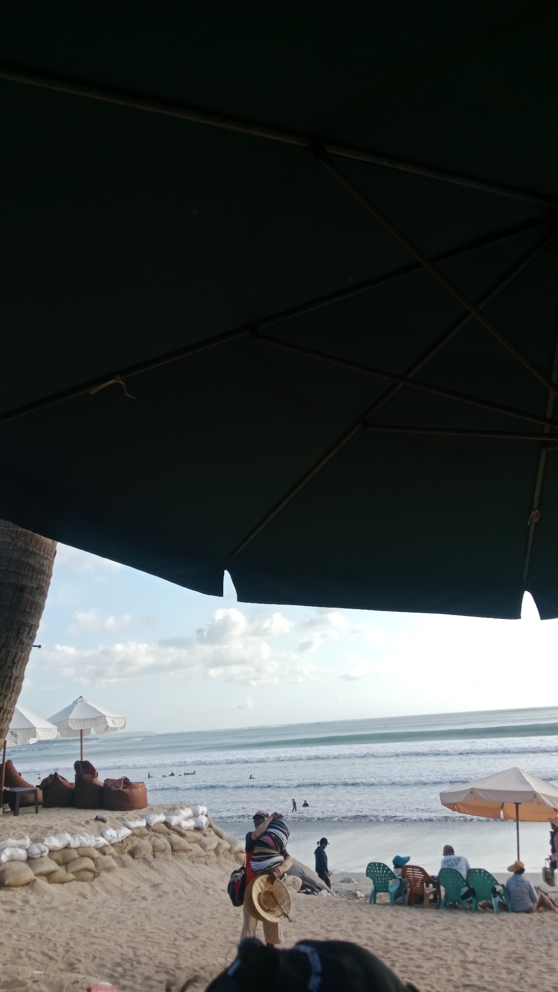 A low-angle, outdoor, medium shot captures a beach scene in Kuta, Indonesia, under a large, dark green umbrella that dominates the upper half of the frame. The umbrella's structural supports are visible against the sky. Below, the sandy beach is populated with various elements. Several large, brown beanbag chairs are arranged on the sand, some behind a row of sandbags. Two white beach umbrellas with the text "LAGOON" are set up further back, near the beanbags. The ocean, with moderate waves and several surfers in the water, stretches across the background under a cloudy sky. In the mid-ground, a person is carrying a straw hat and a colorful bag. Further to the right, a group of people are seated in colorful plastic chairs under a beige umbrella. The scene conveys a relaxed atmosphere typical of a beach resort.