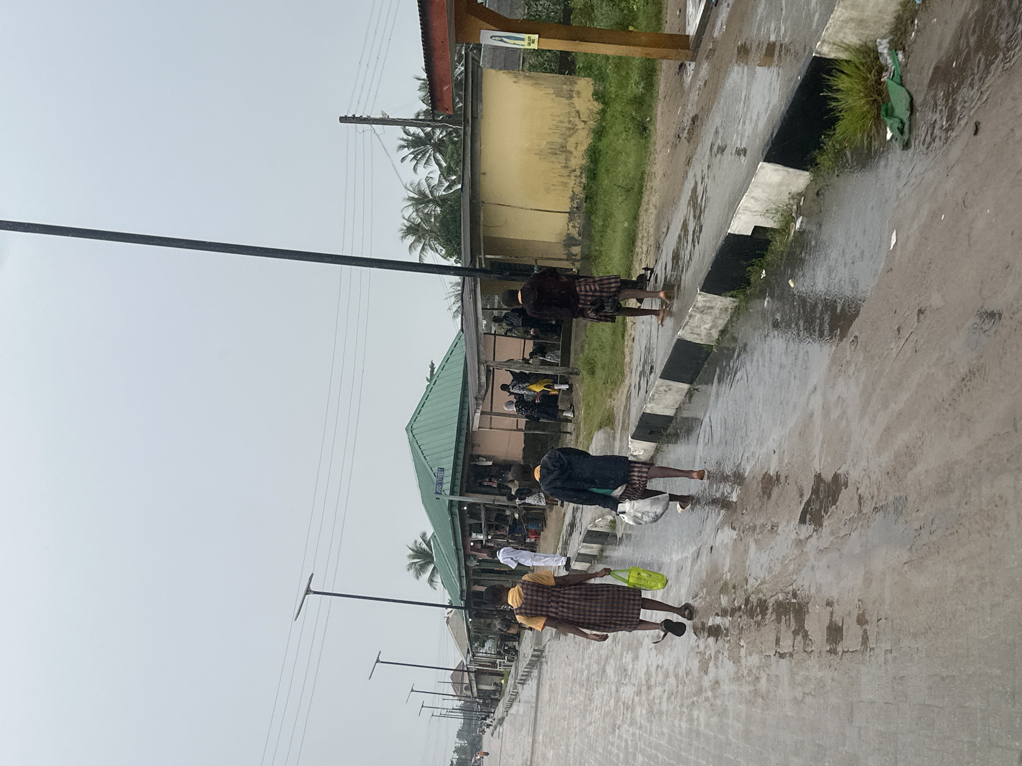 The image depicts a street scene in Badagry, Nigeria, on an overcast day. The street is paved with interlocking bricks and is wet, indicating recent rain or ongoing light precipitation. Several people are visible walking along the street and in front of small shops lining the right side of the road.
In the foreground, two individuals, likely children based on their attire and size, are walking away from the camera. The person on the left wears a checkered brown and yellow dress and carries a bright yellow bag. The person on the right wears a dark hooded jacket and a checkered skirt and carries a white bag. Both are walking on the wet pavement.
Further down the street, several other people are present. One person in a white garment stands near a stall. Others are seen near the shops, some appearing to be customers or vendors. The shops are low-rise structures with corrugated metal roofs, some painted yellow or with green roofs. A sign indicating "AOGI STREET" is visible above one of the green-roofed buildings.
Streetlights with modern, angular designs are spaced along the left side of the road, and power lines are strung overhead. Vegetation, including palm trees, is visible in the background, suggesting a tropical or sub-tropical environment. The overall scene conveys a sense of everyday life in a community with modest infrastructure, experiencing inclement weather.