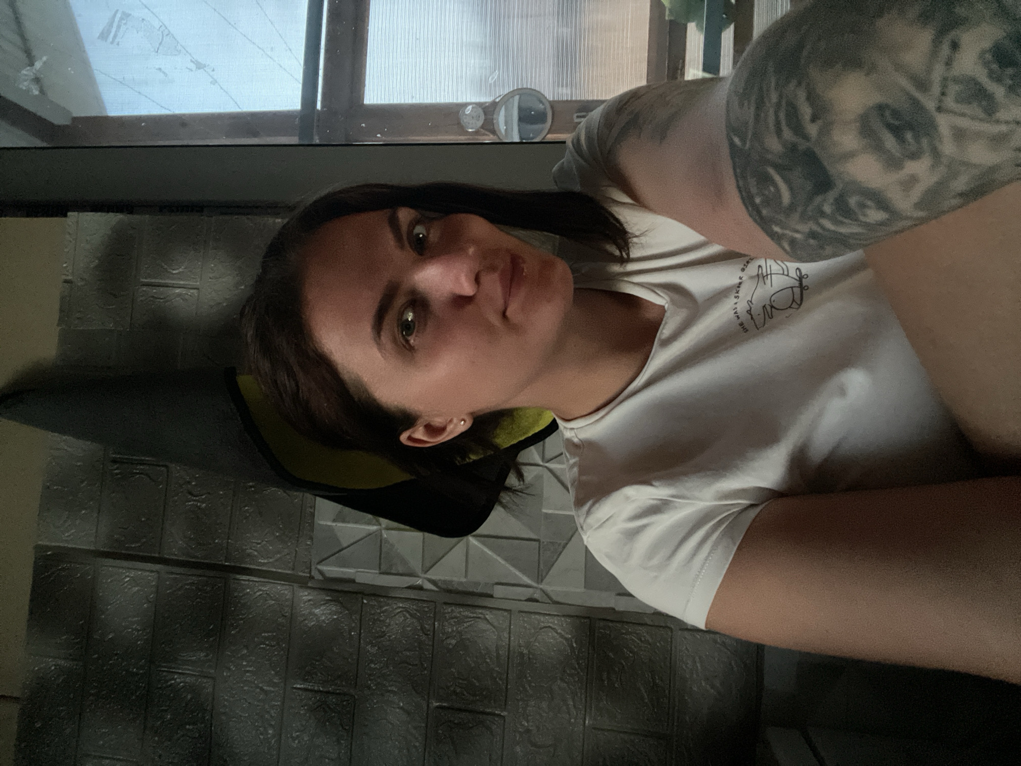 A woman with shoulder-length dark hair, fair skin, and a light-colored t-shirt with a graphic on it is positioned in front of a wall covered in rectangular, patterned tiles. She is looking at the camera. Her right arm, heavily tattooed, is angled across the frame. Behind her, a window with translucent glass panels is visible. The location appears to be indoors, possibly in or near a building with tiled walls and modern windows. The photo was possibly taken in Мосметаллоконструкция Онт, Russia.