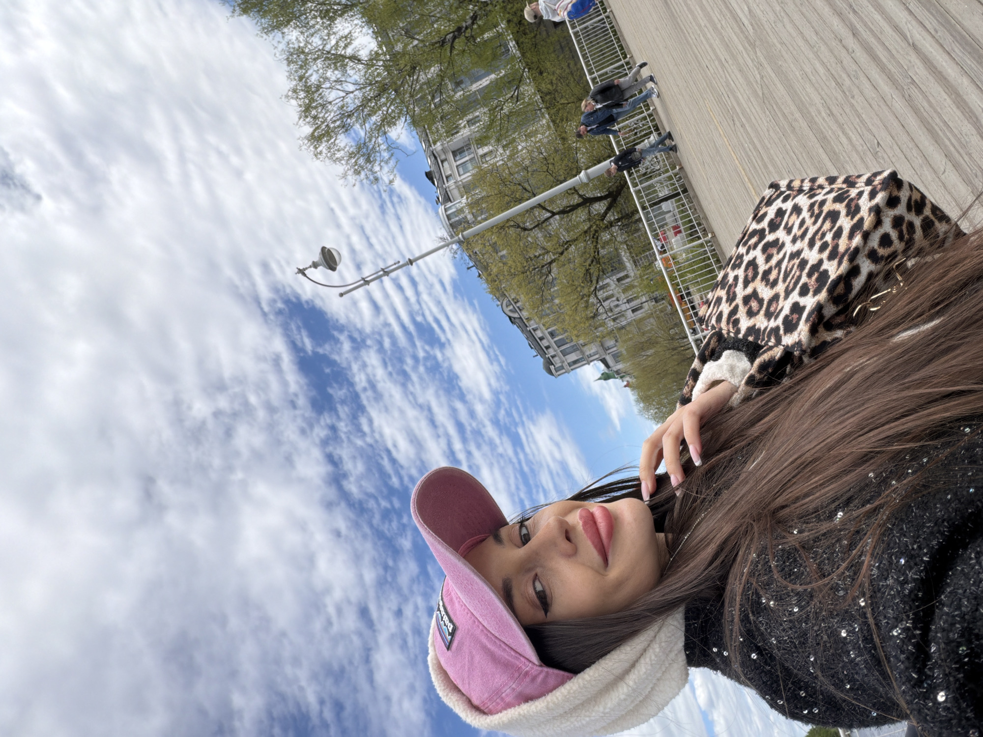 The image depicts a woman in a pink cap and patterned jacket lying down in what appears to be a public space. The background includes a building, trees, a lamppost, and a bridge. The location is identified as Saint Petersburg, Russia based on the combination of architectural elements, the lamppost style, the bridge and foliage common to the city's landscape. Several people can be seen walking on the bridge. The sky is visible with a mixture of blue and white clouds.