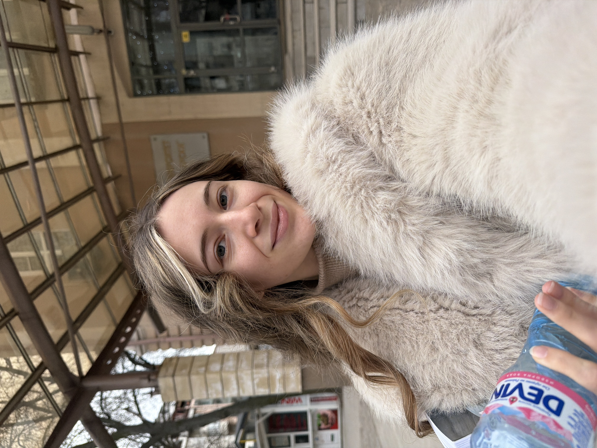 A woman with blonde-streaked brown hair, wearing a light beige faux fur coat, smiles at the camera. She holds a bottle of "Devin" brand water. Behind her is an outdoor structure with brown metal beams and a building with a sign that appears to say "POŞTA", indicating a post office. In the background, bare trees are visible, along with a small white kiosk-like structure, suggesting an urban environment. This location is identified as Plovdiv, Bulgaria, based on the "POŞTA" sign (likely related to the Bulgarian postal service) and the water bottle brand.