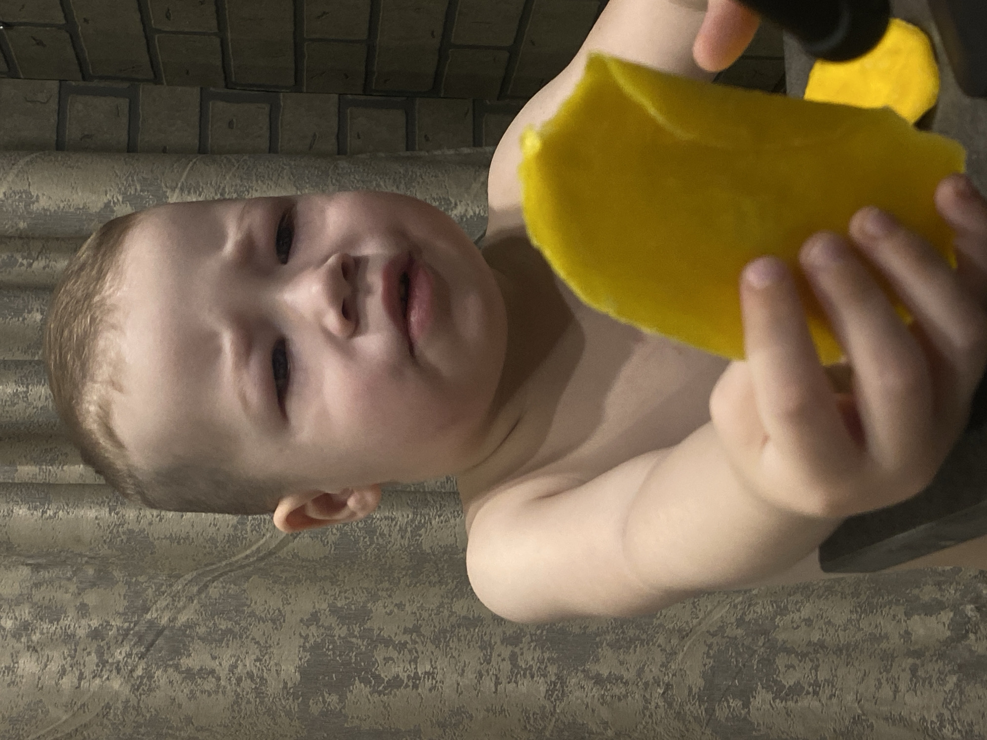 A child is holding a yellow sponge. The child has fair skin, light brown hair, and is shirtless. The background is a gray wall with a textured pattern. The location in the city of Sochi, Russia, cannot be determined from the image.