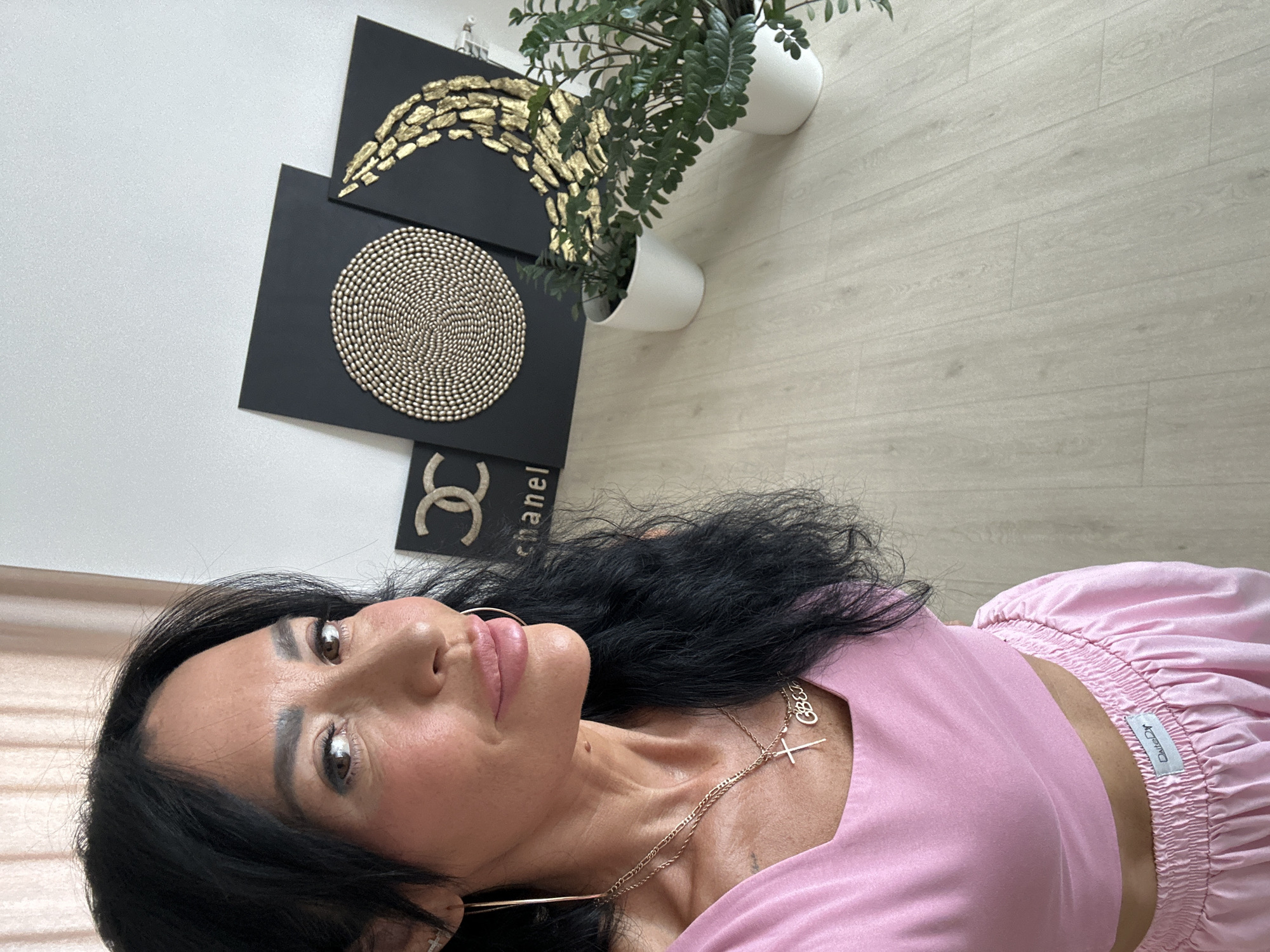 A woman with dark hair and brown eyes is taking a selfie in a room. She is wearing a pink crop top and pink bottoms. She has on gold hoop earrings and two necklaces, one with a cross and another with a charm. In the background, there are three black framed art pieces with gold designs and two potted plants on a light-colored wooden floor. The room has a light-colored wall and a curtain in the background. There is no indication of the city of Tyumen, Russia.