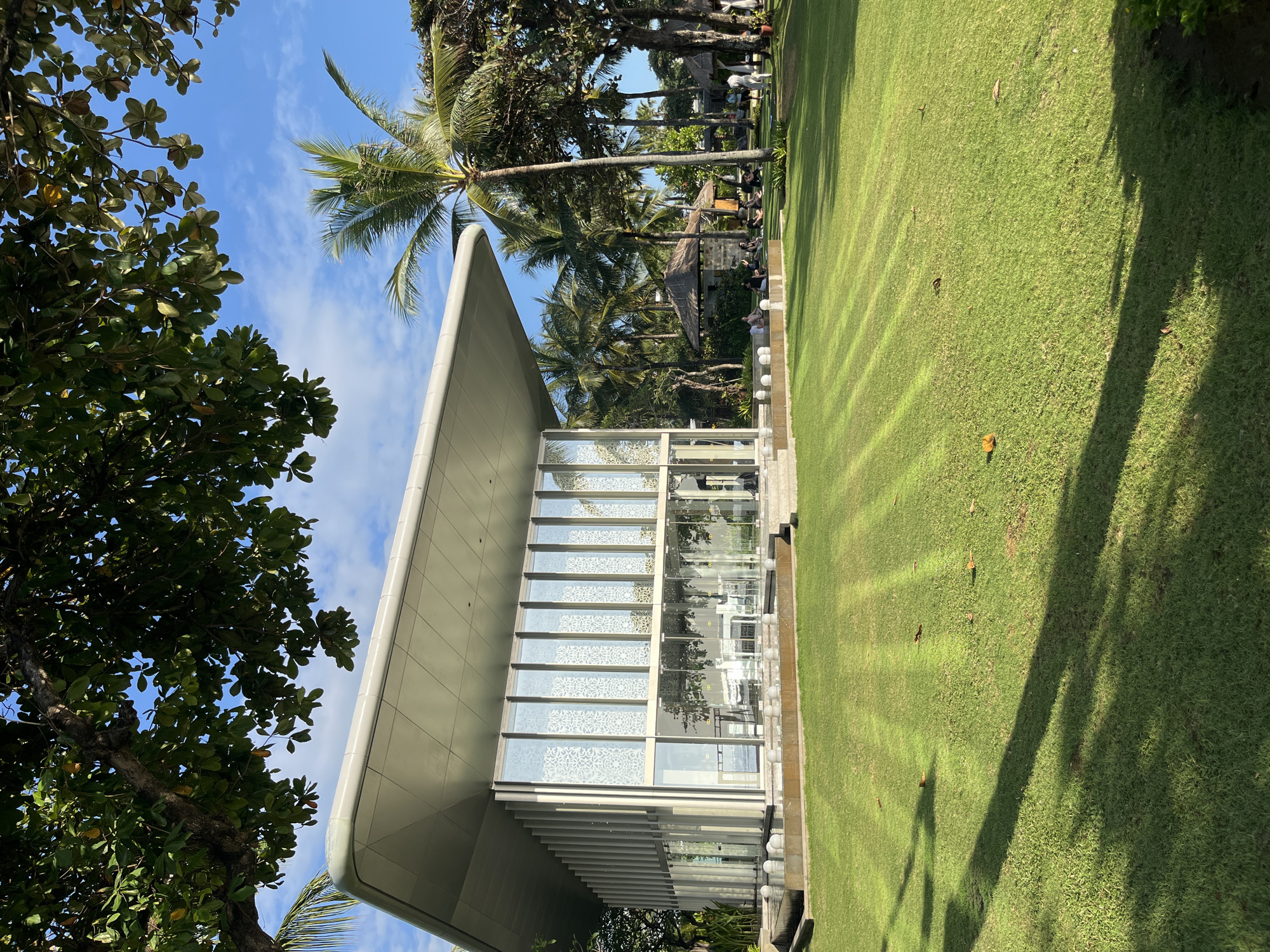 This image, taken in Kuta Selatan, Indonesia, depicts a modern, glass-walled building with a cantilevered roof set on a well-manicured lawn. The building has a unique facade with vertical fins and decorative patterned glass panels. In front of the building, a shallow water feature with spherical elements adds a decorative touch. The lawn is illuminated by sunlight, creating distinct striped patterns from the mowing. Scattered across the lawn are a few fallen leaves. In the background, lush tropical foliage, including palm trees, and several thatched-roof structures are visible. A group of people are gathered in the background, some sitting and others appearing to be engaged in an activity. The sky is blue with scattered white clouds.