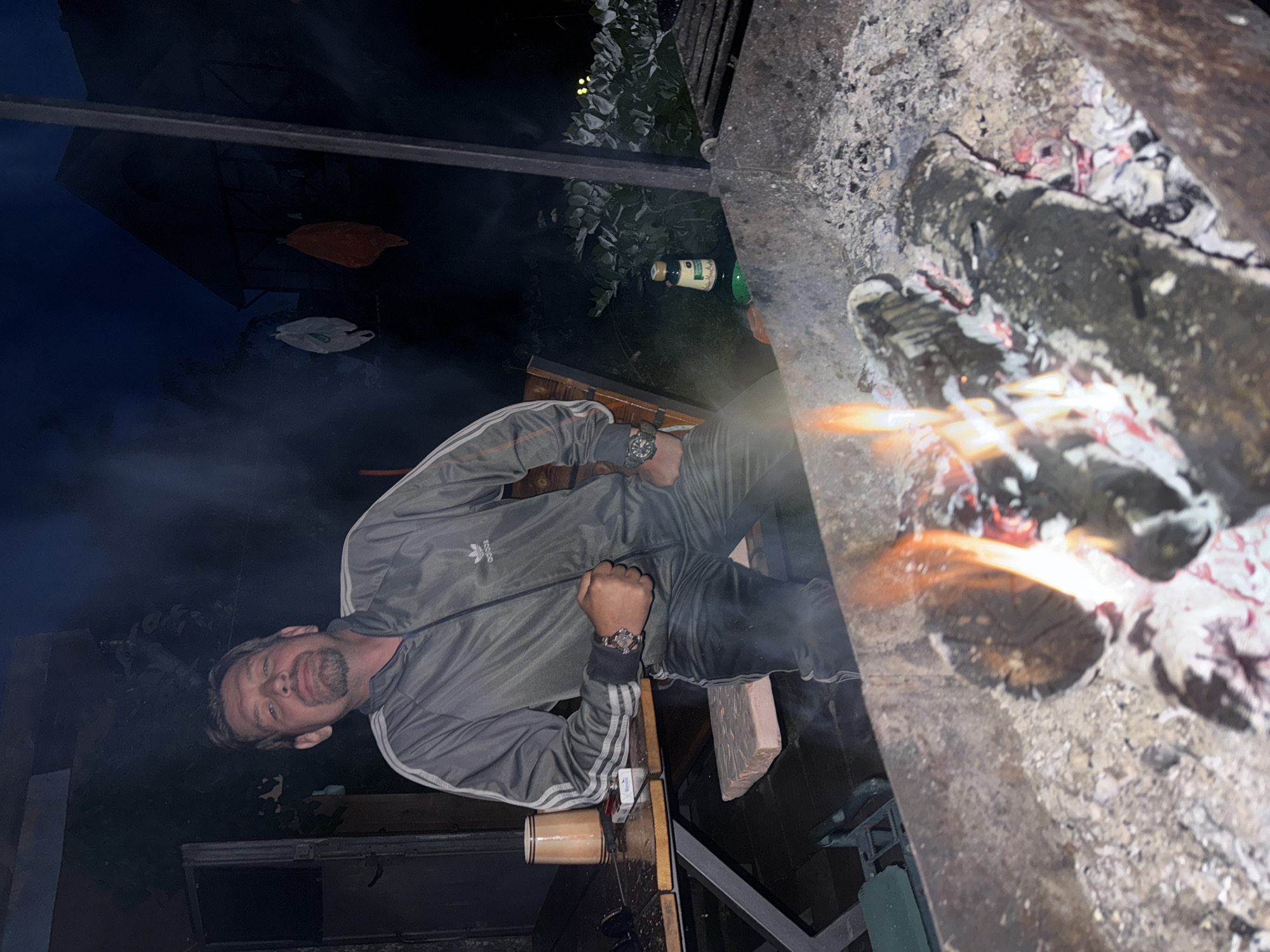 A man wearing a grey Adidas tracksuit stands next to a barbecue pit at dusk. He has short, dark hair and a goatee, and is looking at the camera with his left hand resting on his chest. A cigarette pack and a mug are on a wooden table to his left. The barbecue pit in the foreground is filled with glowing embers and burning logs. In the background, a building with lights on is visible, along with trees and the night sky. The location is likely a backyard or outdoor recreational area in Автомобилист-1 Снт, Russia, given the presence of the barbecue and the general ambiance.