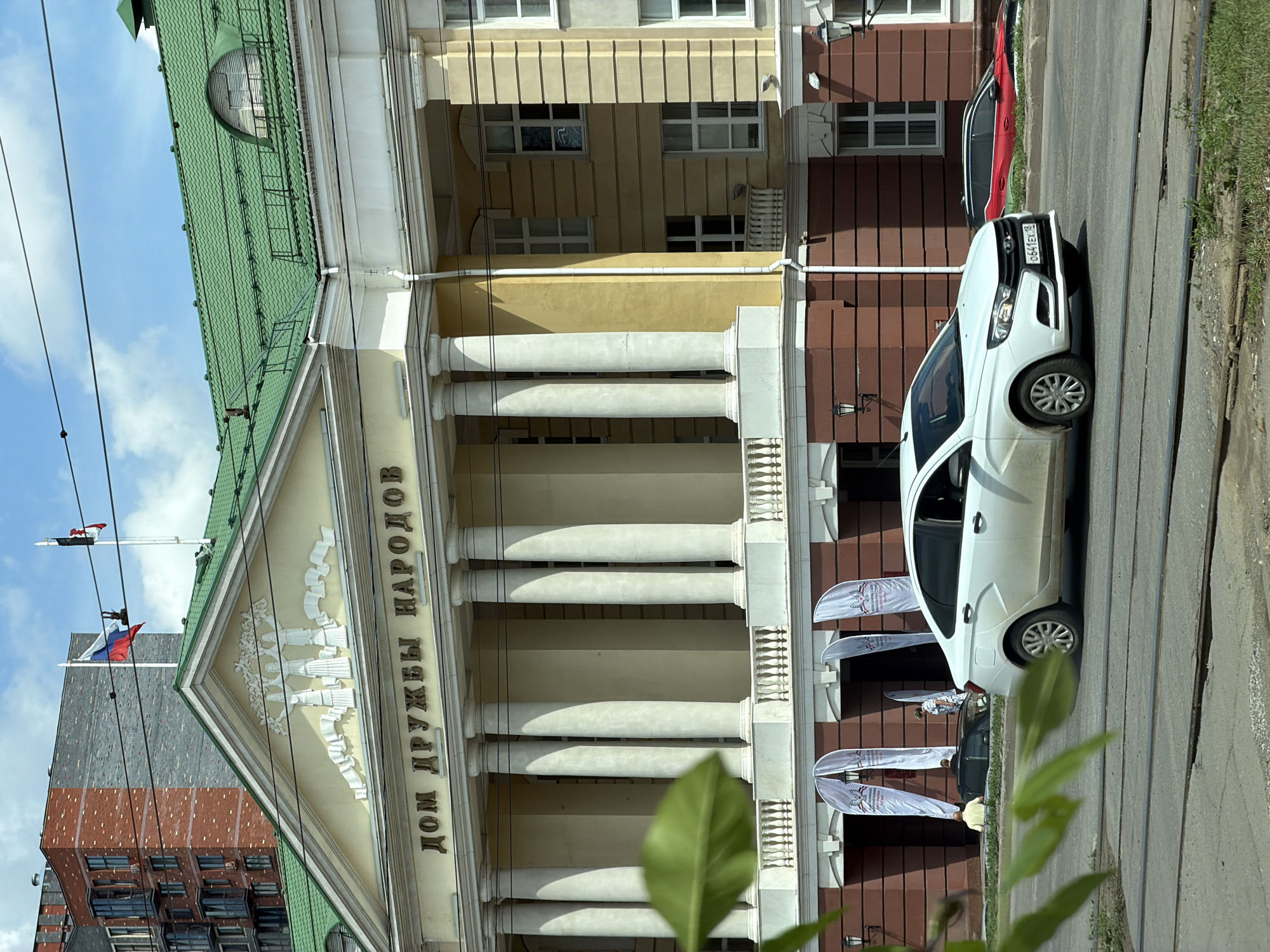 An exterior, eye-level shot captures the House of Friendship of Peoples in Izhevsk, Russia, on a sunny day. The building is a classical structure with a yellow facade, featuring a prominent colonnade of tall, white columns and a green, tiled roof. Above the columns, the building's name is inscribed in large, gold letters, reading "ДОМ ДРУЖБЫ НАРОДОВ" (House of Friendship of Peoples). A bas-relief sculpture depicting figures holding hands adorns the pediment above the inscription.
In front of the building, several white, arch-shaped banners are displayed, bearing some insignia. Two cars are visible on the road in the foreground. A white Lada sedan is driving in the right lane, and a red and black car is parked or driving in the left lane further down the road. A person in a yellow shirt and another in a white dress are visible near the entrance of the building, possibly attending an event. The foreground also shows tram tracks and some green foliage. In the background, a modern brick and glass apartment building stands tall, contrasting with the classical architecture of the House of Friendship of Peoples. The sky is a clear blue with some scattered clouds.