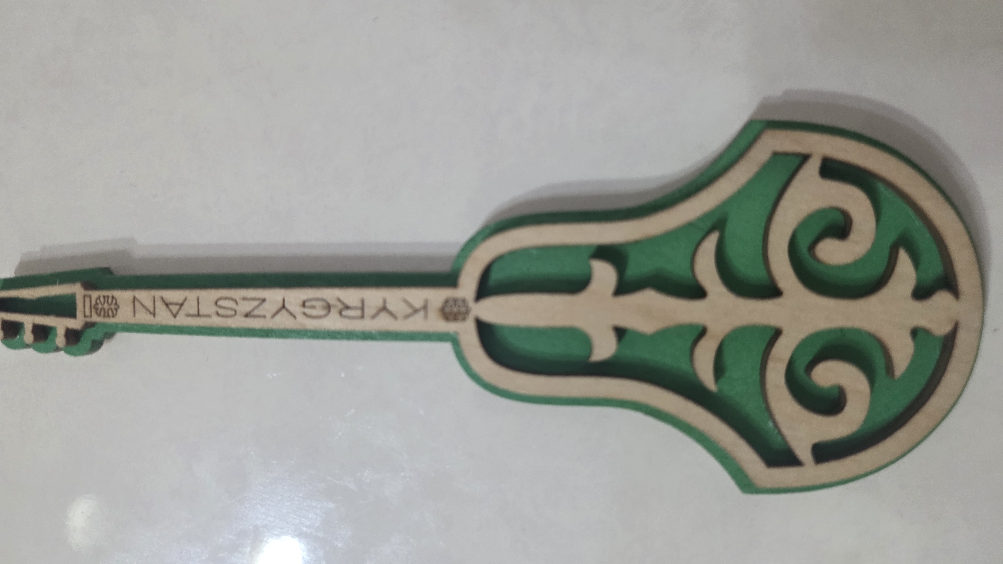 A souvenir in the shape of a musical instrument, possibly a komuz, is displayed against a white background. The souvenir is made of layered wood and painted green. It features decorative cutouts and the word "KYRGYZSTAN" inscribed vertically along its neck. The location in Chuy, Kyrgyzstan is not discernible from the image.