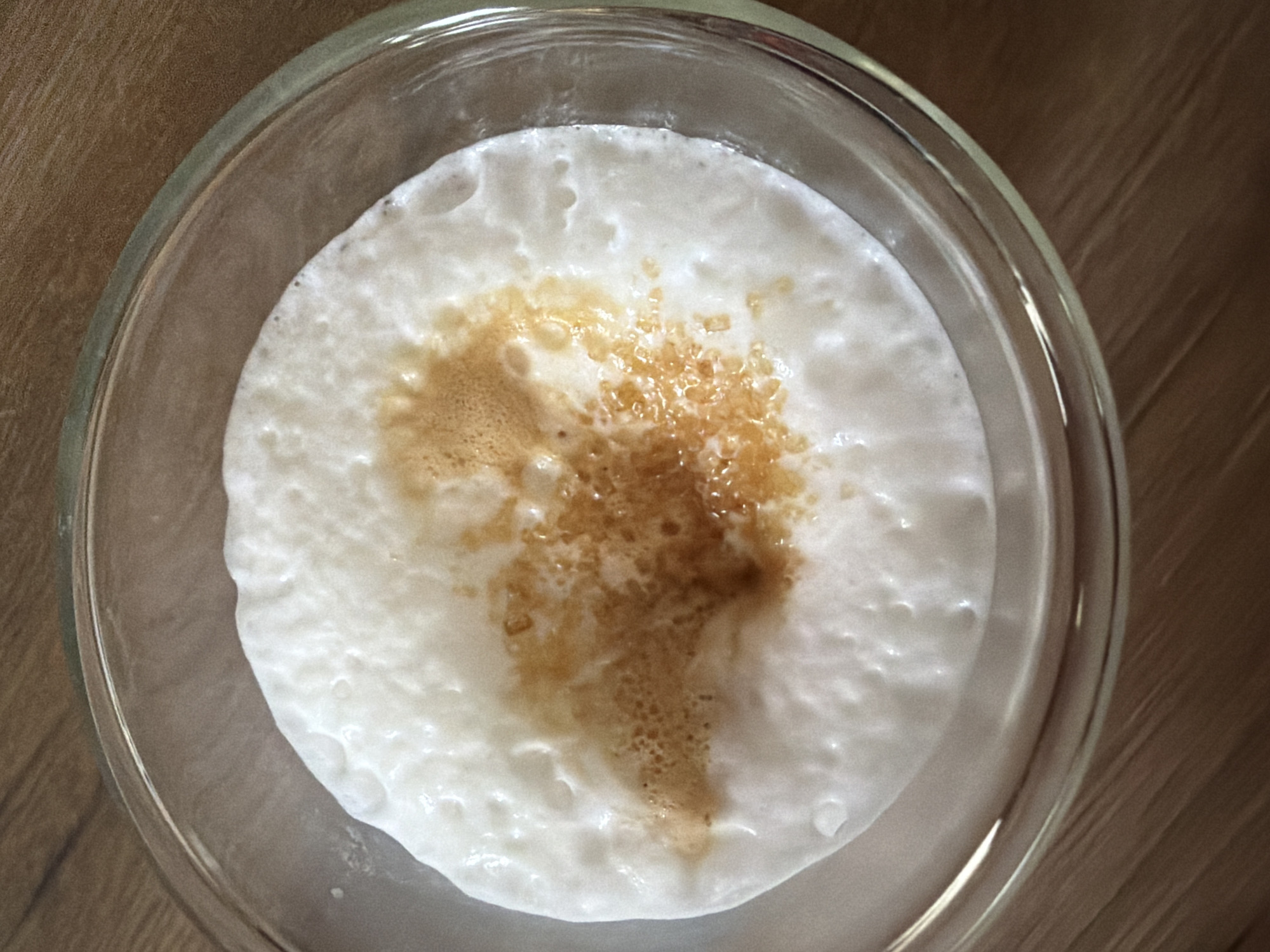 A close-up, overhead shot shows a clear glass bowl containing a white, foamy substance, likely milk or cream, topped with a light brown, granular substance that appears to be sugar. The sugar is concentrated in the center of the bowl, with some of it having mixed with the foam, creating streaks of brown. The bowl is placed on a wooden surface with a visible grain pattern. The lighting suggests an indoor setting. There is no discernible connection to Pazardzhik, Bulgaria, or any specific environmental context that would place it in a city. The image is purely a still-life of a food or beverage preparation.