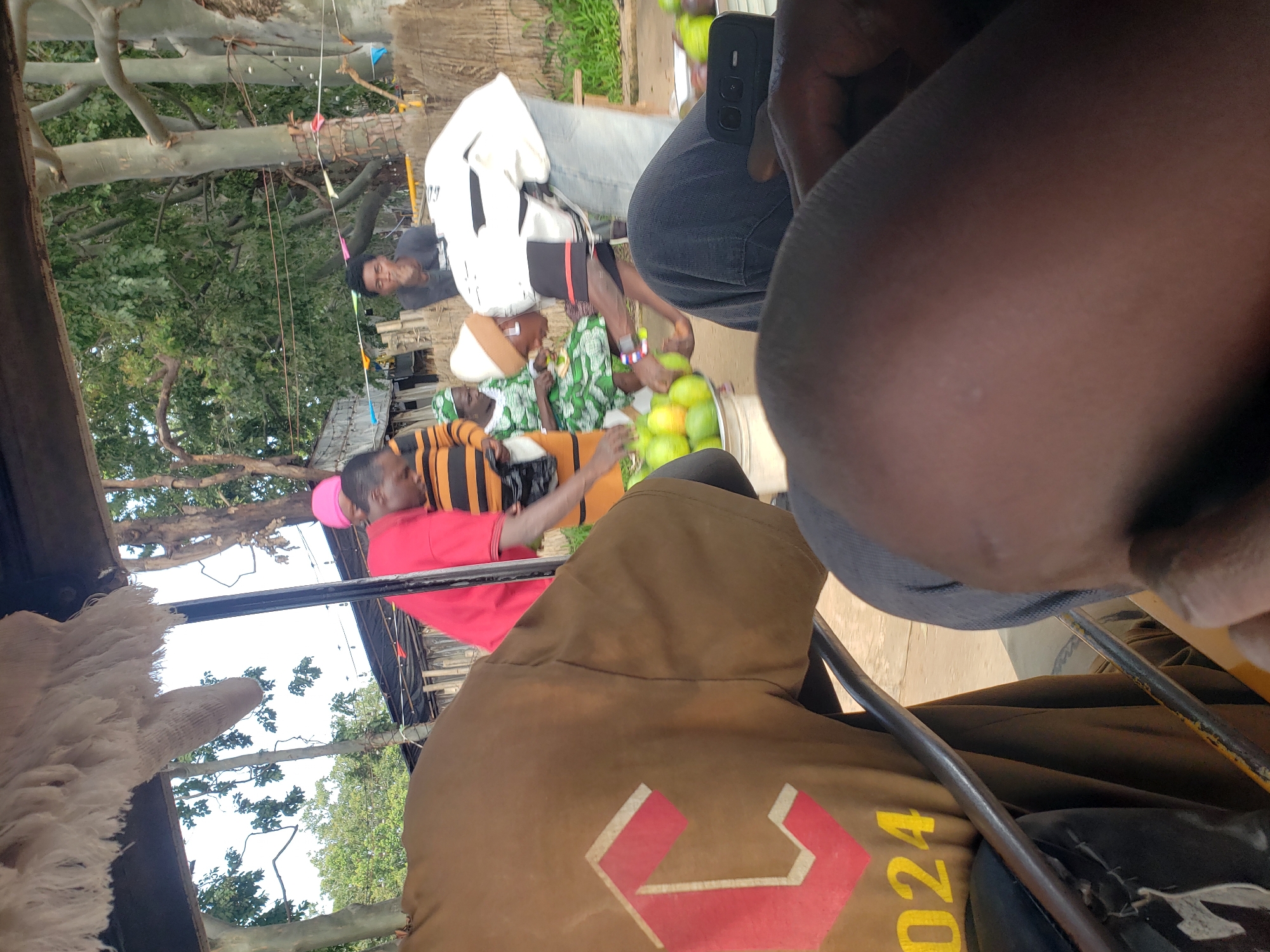 A street scene in Gura Top, Nigeria features a group of people under a makeshift canopy. In the foreground, a person wearing a brown t-shirt with a red and white "C" logo and "024" printed in yellow is seated. To the right, another person's legs are visible, with a black mobile phone resting on their lap. In the midground, a person in a red t-shirt stands near another person wearing a black and orange striped shirt. Further back, a woman in a green patterned dress and white headwrap is handing fruit, possibly mangoes, to another individual. Several mangoes are visible in a bucket. The environment appears to be outdoors with trees and some structures in the background.