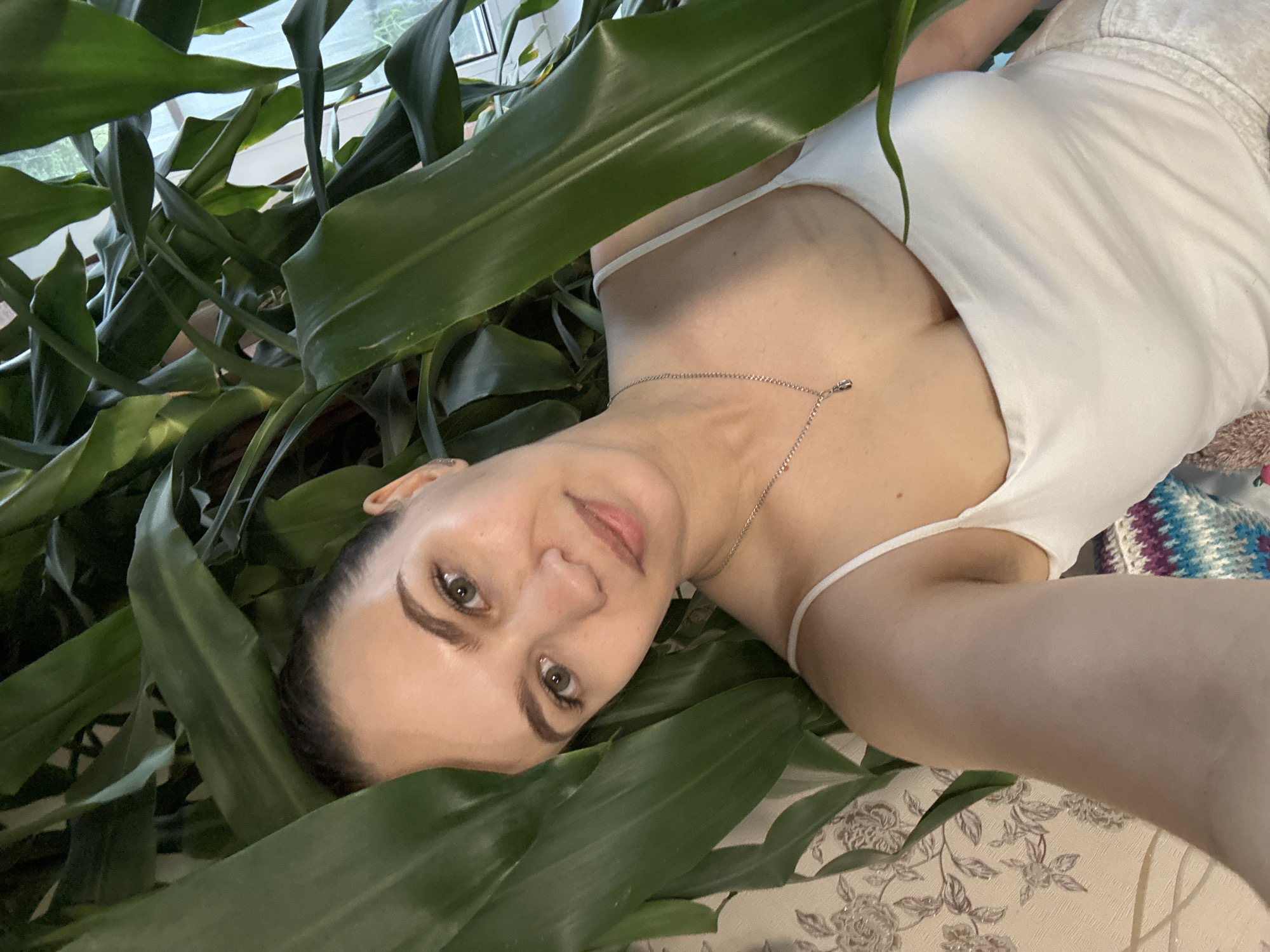 A woman lies among large green leaves, possibly houseplants. She is fair-skinned with dark hair pulled back, wearing a white tank top and light-colored shorts. She has a silver necklace. The background includes floral patterned wallpaper and possibly a knitted blanket. The location, Nizhnyaya Gar', Russia, cannot be determined from the image alone.