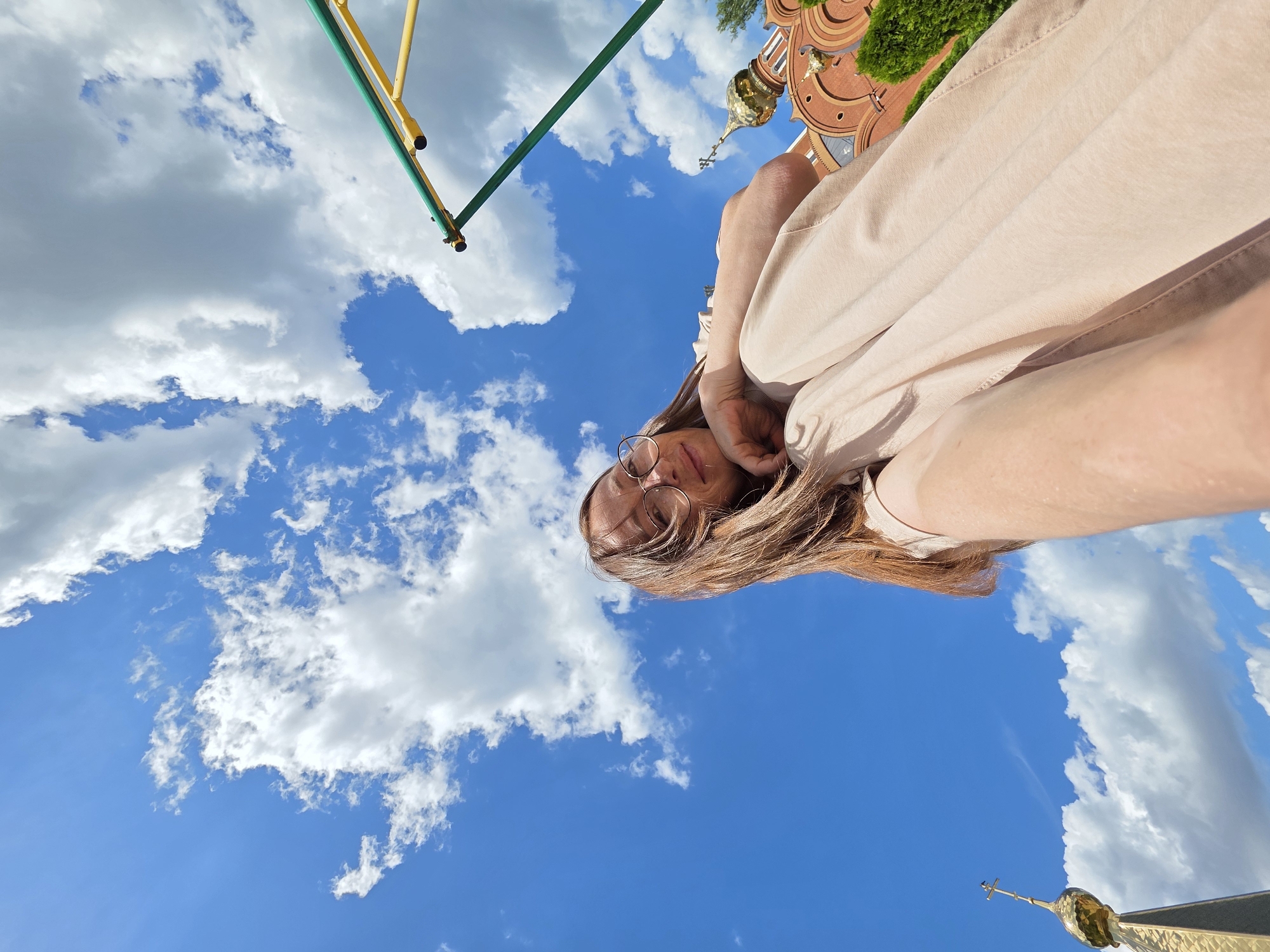 The image shows a young woman with long brown hair and glasses, wearing a light-colored outfit, sitting in front of a building with golden domes, possibly a church. The building appears to be red brick with arched windows. The sky is blue with scattered white clouds, and a portion of a playground structure with green and yellow bars is visible in the upper right corner. The perspective is from a low angle, looking up at the woman and the sky. The photo seems to have been taken outdoors on a sunny day. The building's architecture suggests it might be located in Russia, and given the prompt's context, it could be in the city of Chaikovsky.