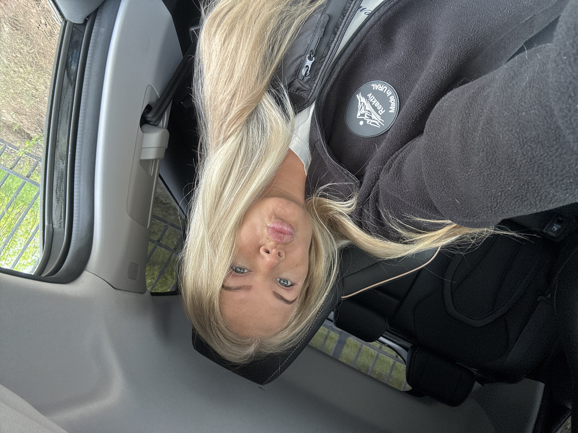 A fair-skinned, blonde woman is inside a car, turned sideways to the camera. She has blue eyes and is pursing her lips in a "duck face." She wears a black fleece jacket with a "Reaktiv made in URAL" patch. The car has gray interior and black leather seats. Outside the car window is vegetation and a dark-colored metal fence. It is impossible to definitively place the scene in Chaikovsky, Russia without further information or context.