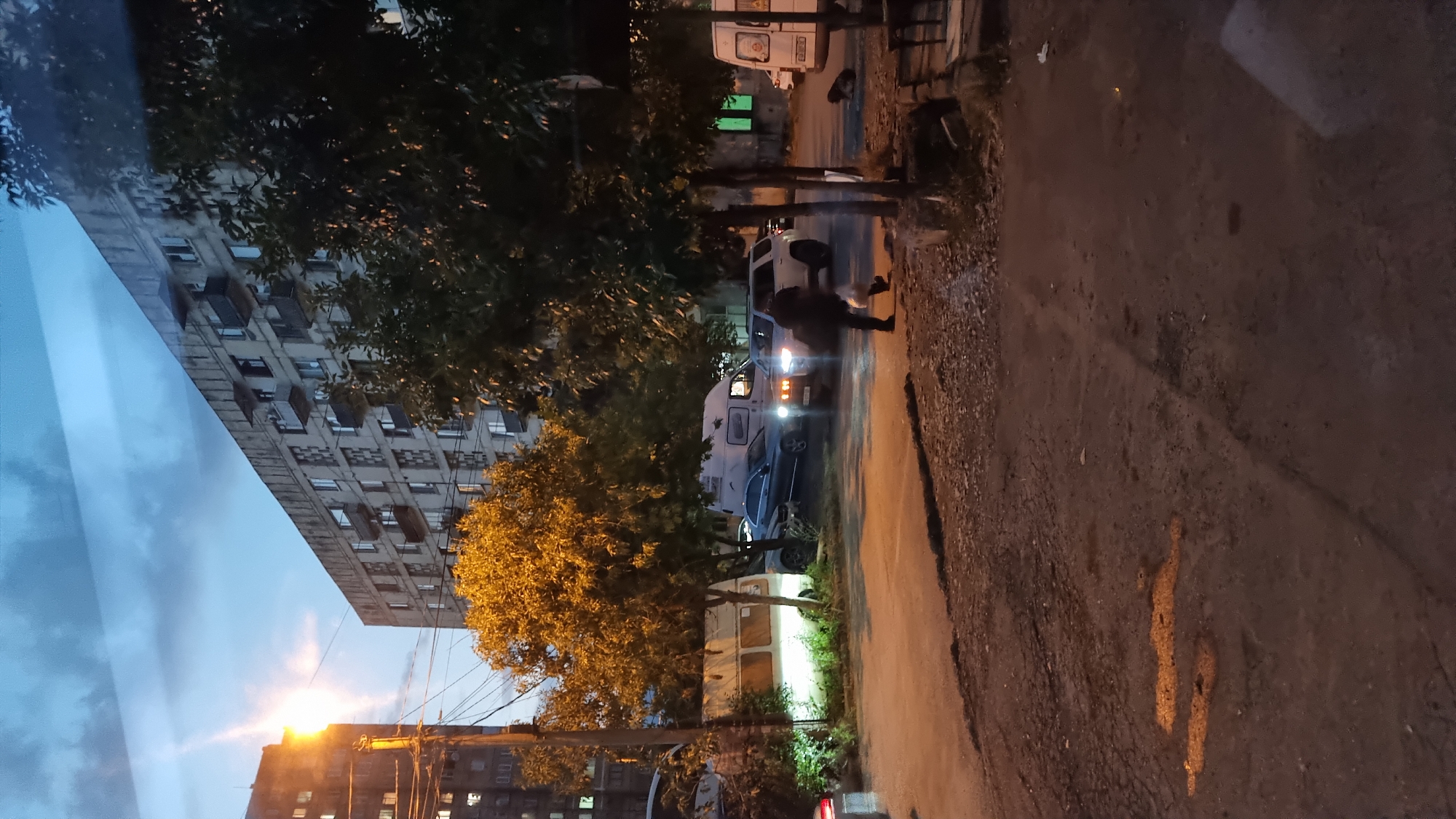 The image depicts an evening scene on a street in Tbilisi, Georgia, with residential buildings lining the background and trees in the foreground. A person is walking across the street, carrying what appears to be a plastic bag. Several vehicles are present, including a silver SUV, a blue sedan, and two white vans, one of which has its headlights on. One of the vans is parked near a tree. The street surface shows signs of disrepair, with visible cracks and uneven patches. The sky is dark, indicating it is either dusk or night, with some clouds visible. A streetlight on the left side of the image is illuminated. The overall atmosphere suggests a typical urban setting in Tbilisi during the evening.