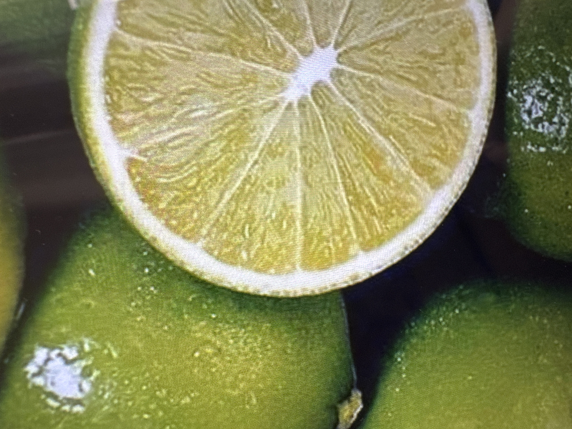 The image is a close-up, vertically oriented photograph of several limes. A large portion of the image is filled with the cut surface of a lime, revealing its segmented interior and white pith. To the left of the cut lime, the rind of another whole lime is visible, showing a slightly textured, glossy green surface with some water droplets. The background is dark and out of focus, suggesting a shallow depth of field. There are no people or any identifiable elements of the city of Yalta, Ukraine, in the image.