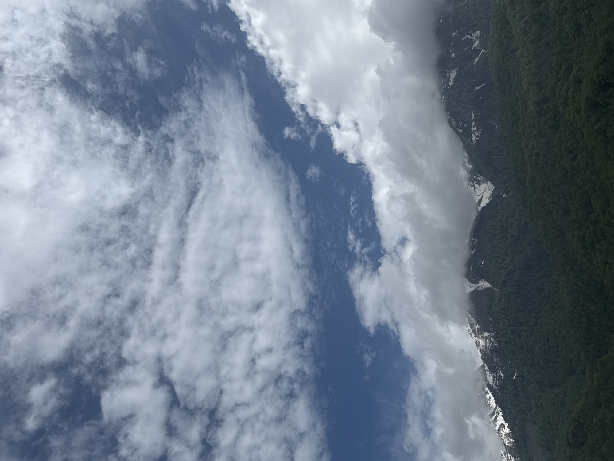 The image shows a landscape scene. On the right side of the frame, a mountain range covered in dense, green vegetation is visible. Patches of snow and grey rock formations are scattered along the mountain's steep slopes. Adjacent to the mountains, thick white clouds partially obscure the view. The left side of the image is dominated by a vast blue sky with numerous white, fluffy clouds. The perspective of the photo suggests the view is tilted upward, possibly taken from a lower altitude looking towards the sky and mountains.
The location in the city of Krasnaya Polyana, Russia cannot be determined.