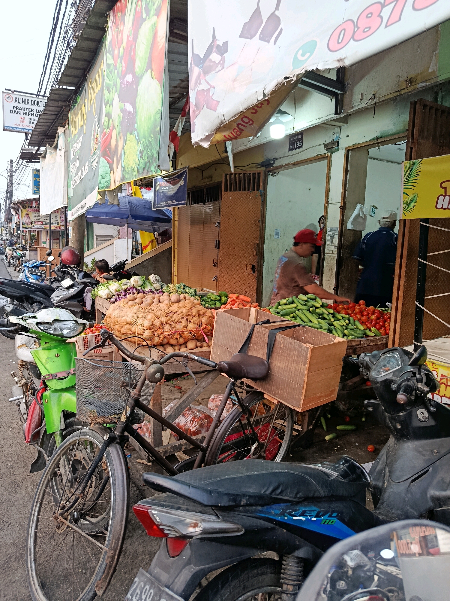 The image depicts an outdoor market scene in the city of Bekasi, Indonesia, characterized by a street-level view of various stalls and parked motorcycles. Prominently displayed are large banners advertising businesses, one of which features images of vegetables. In the foreground, a black bicycle with a wire basket is parked next to a green motorcycle. The bicycle's rear wheel and part of its frame are visible. Behind the bicycle, a variety of fresh produce is arranged on display, including a large sack of potatoes, piles of cucumbers and tomatoes, and some leafy vegetables. Two wooden crates are filled with more vegetables. A man wearing a red cap is seen interacting with the produce, likely a vendor. In the background, several motorcycles are parked along the street, and doorways of shops are visible. A few people are present in the background, including one man wearing a dark blue shirt and a cap, and another person partially obscured. The overall environment suggests a bustling marketplace with an emphasis on fresh produce sales.