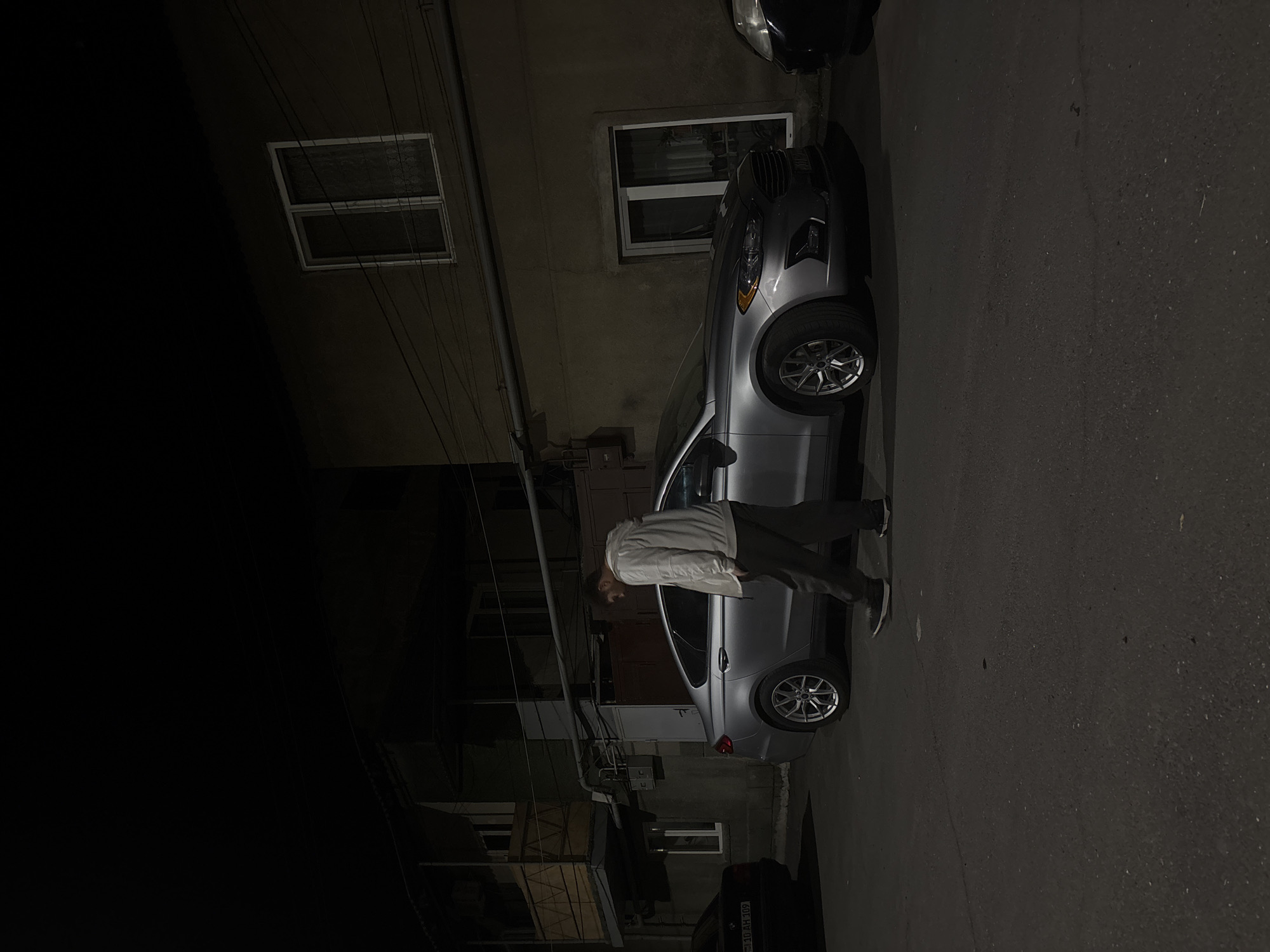 The image, taken at night in what appears to be a residential area of Yerevan, Armenia, depicts a silver car parked on the side of a street. The license plate of the car parked behind it has visible letters. A person is standing next to the silver car. Buildings with windows are visible in the background. Electrical lines are strung overhead. The asphalt street shows signs of wear.