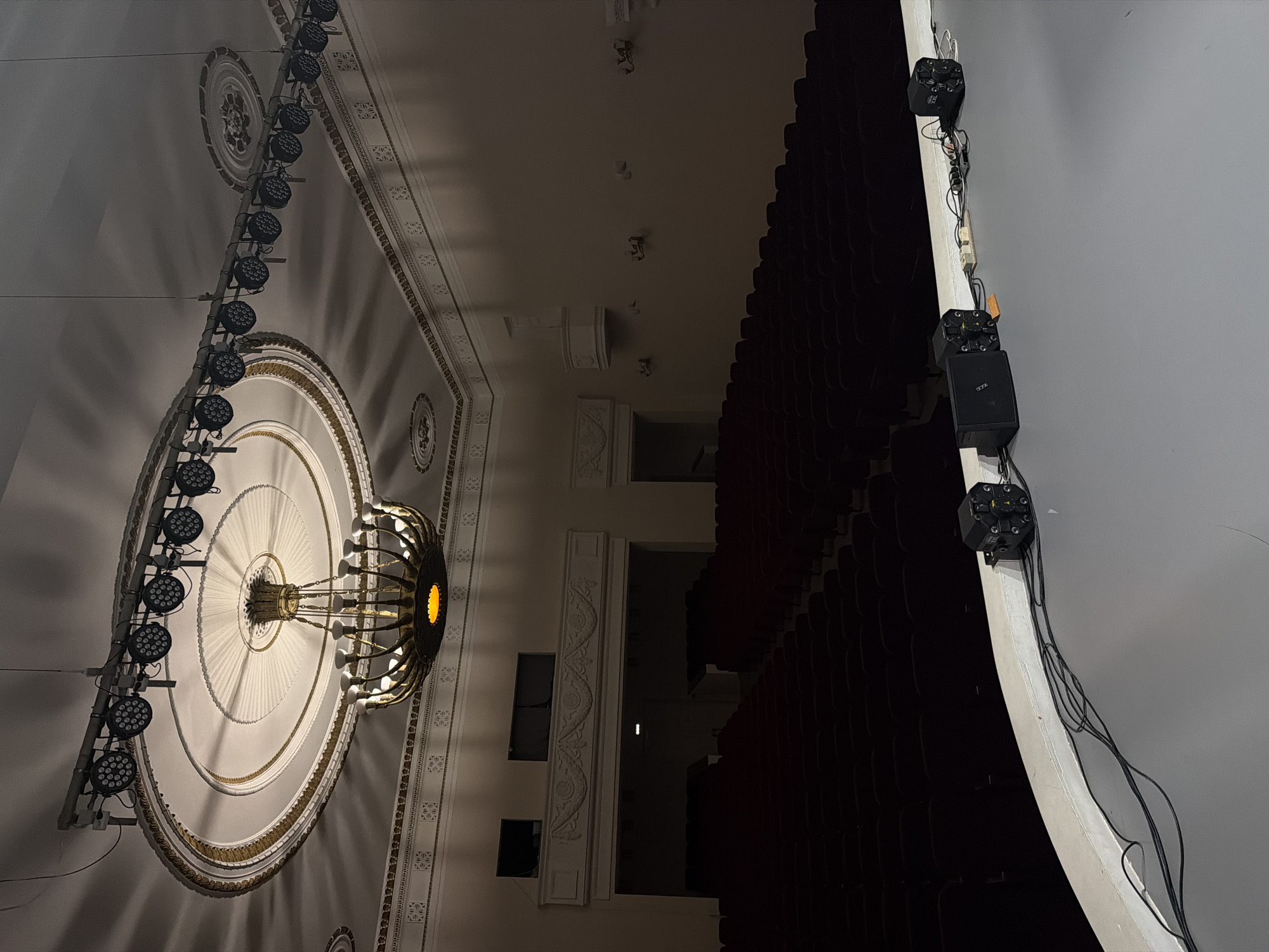 The image shows the interior of a theater, possibly in Perm, Russia. The focus is on the ornate ceiling featuring a large chandelier with several lights and a circular, decorative molding. A row of lighting fixtures is mounted on a track alongside the decorative ceiling element. The theater's seating area is visible on the right side, with rows of seats extending into the darkness. On the far right, black boxes attached to a beam are wired to each other. The color palette is dominated by dark shadows and dim lighting.
