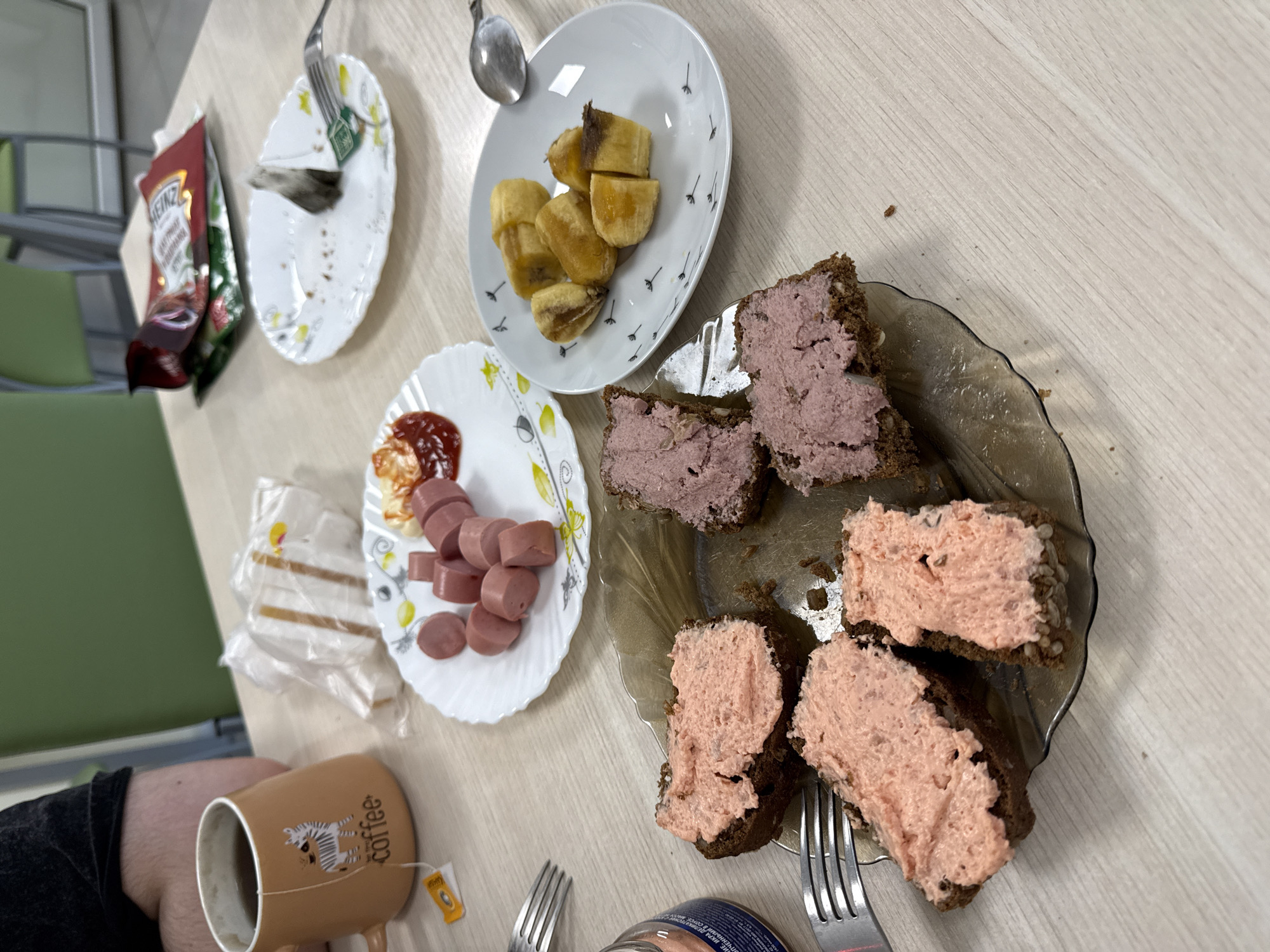 The image depicts a table setting with various food items, likely in Moscow, Russia. There are multiple plates: one with what appears to be fried bananas, another with sliced sausages and a dollop of ketchup, and a third with several open-faced sandwiches topped with a pinkish-purple spread. A mug of tea is present, as is a packet of Heinz ketchup. Cutlery (forks and a spoon) is scattered around the plates. A glimpse of an individual's arm and the edge of a chair is visible. The setting seems to be indoors, possibly a cafeteria or a simple dining area.