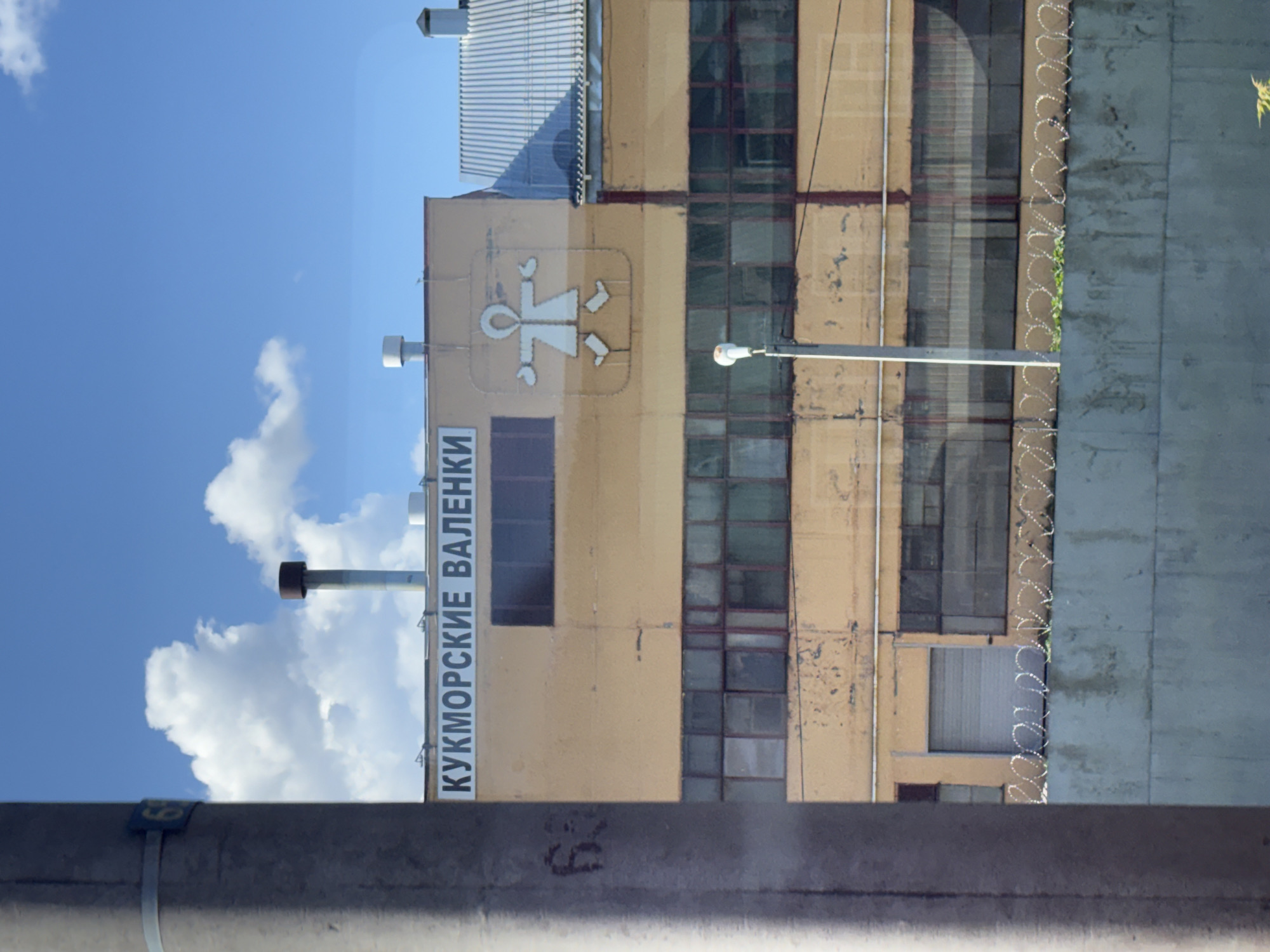 The image presents a static, daytime exterior view of a multi-story building in Kukmor, Russia. The building's facade is primarily a faded yellow-ochre color, exhibiting visible signs of age, wear, and staining. A prominent black text sign, "КУКМОРСКИЕ ВАЛЕНКИ," which translates to "Kukmor Valenki" (Kukmor Felt Boots), is affixed above the upper windows. To the right of this text, a large white stylized logo depicts an anthropomorphic figure with outstretched arms and distinct valenki (traditional Russian felt boots) for feet, set against a lighter rectangular background on the building.
The roof of the main structure features a dark brown/black chimney stack near the center and a white vent pipe to its right. Below the main sign, dark rectangular windows are present. The lower section of the building contains a continuous band of numerous rectangular windows with dark frames, some featuring vertical red elements, reflecting portions of the sky. A light grey roll-up garage door is visible on the lower left portion of the facade. An adjacent or attached structure with a corrugated metal roof is partially visible to the right of the main building.
In the immediate foreground on the left, a large grey utility pole partially obstructs the view. A small blue rectangular sign bearing the number "63" is attached to this pole. Across the mid-ground, a light grey concrete wall is visible, surmounted by multiple strands of coiled barbed wire. Sparse green vegetation grows immediately behind the barbed wire. A white street light on a grey pole rises from behind the concrete wall on the right side of the frame. The background consists of a clear blue sky with scattered white cumulus clouds. No individuals are visible in the scene.