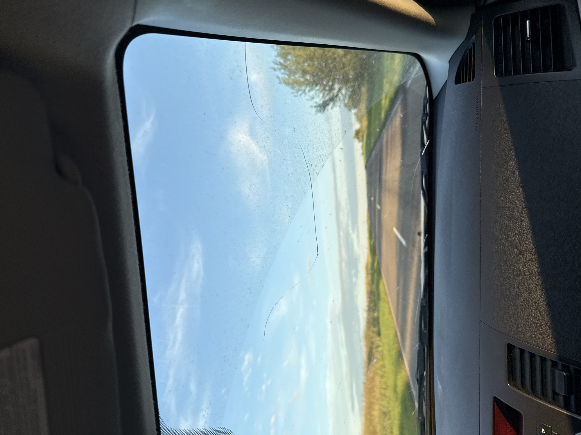 This image is a first-person perspective from inside a vehicle, looking through the windshield. The windshield is dirty and has a visible crack running across it. Outside, a paved road stretches into the distance, flanked by grassy fields and trees under a partly cloudy blue sky. The sun appears to be low in the sky, casting long shadows and illuminating the scene with warm light. The dashboard of the car is visible in the foreground, with a portion of the center console showing a red display and air vents. There are no people or animals visible in the image. The location is identified as Dzhirim, Russia.